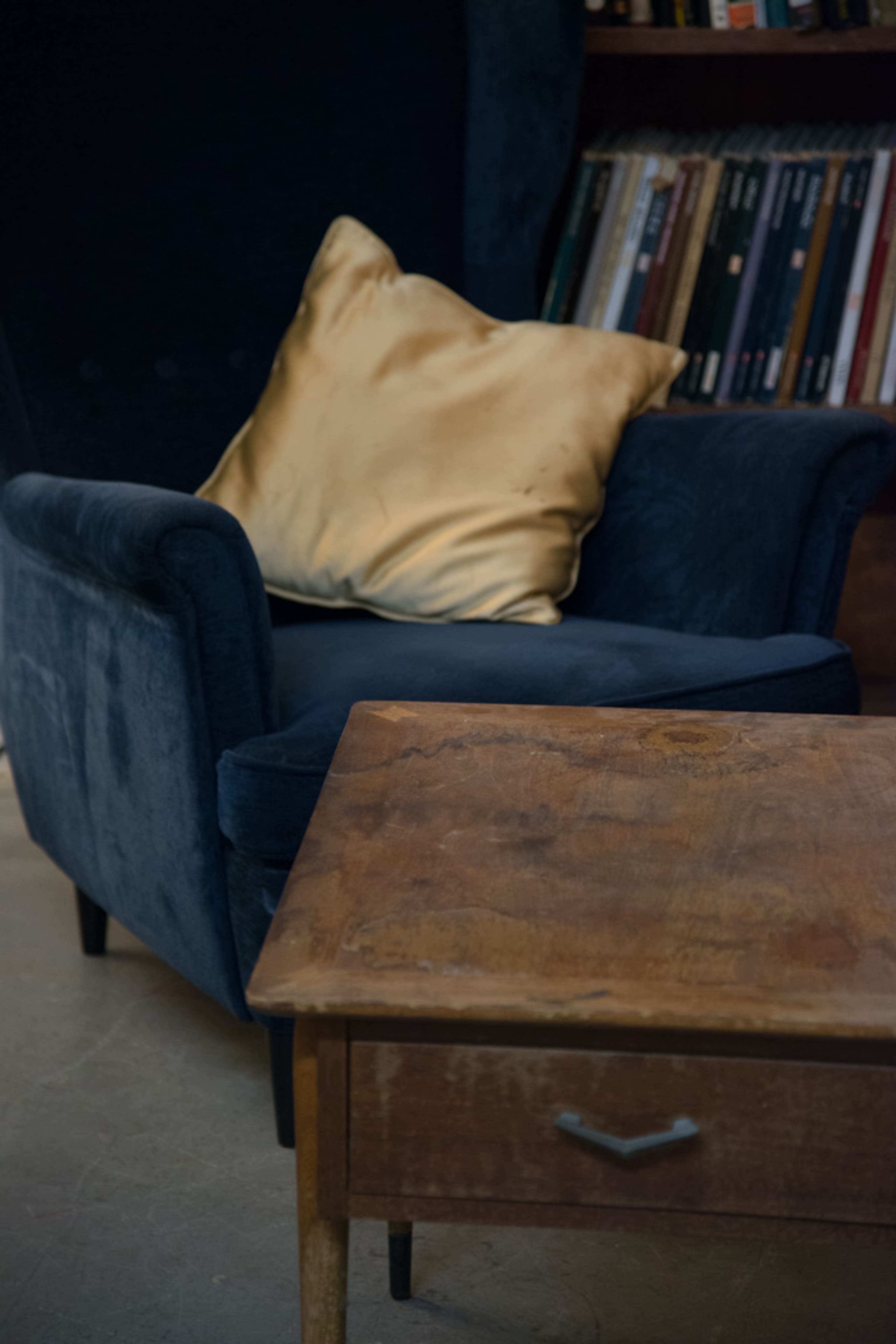 A blue armchair with a gold pillow is positioned next to a wooden table in a room with bookshelves.