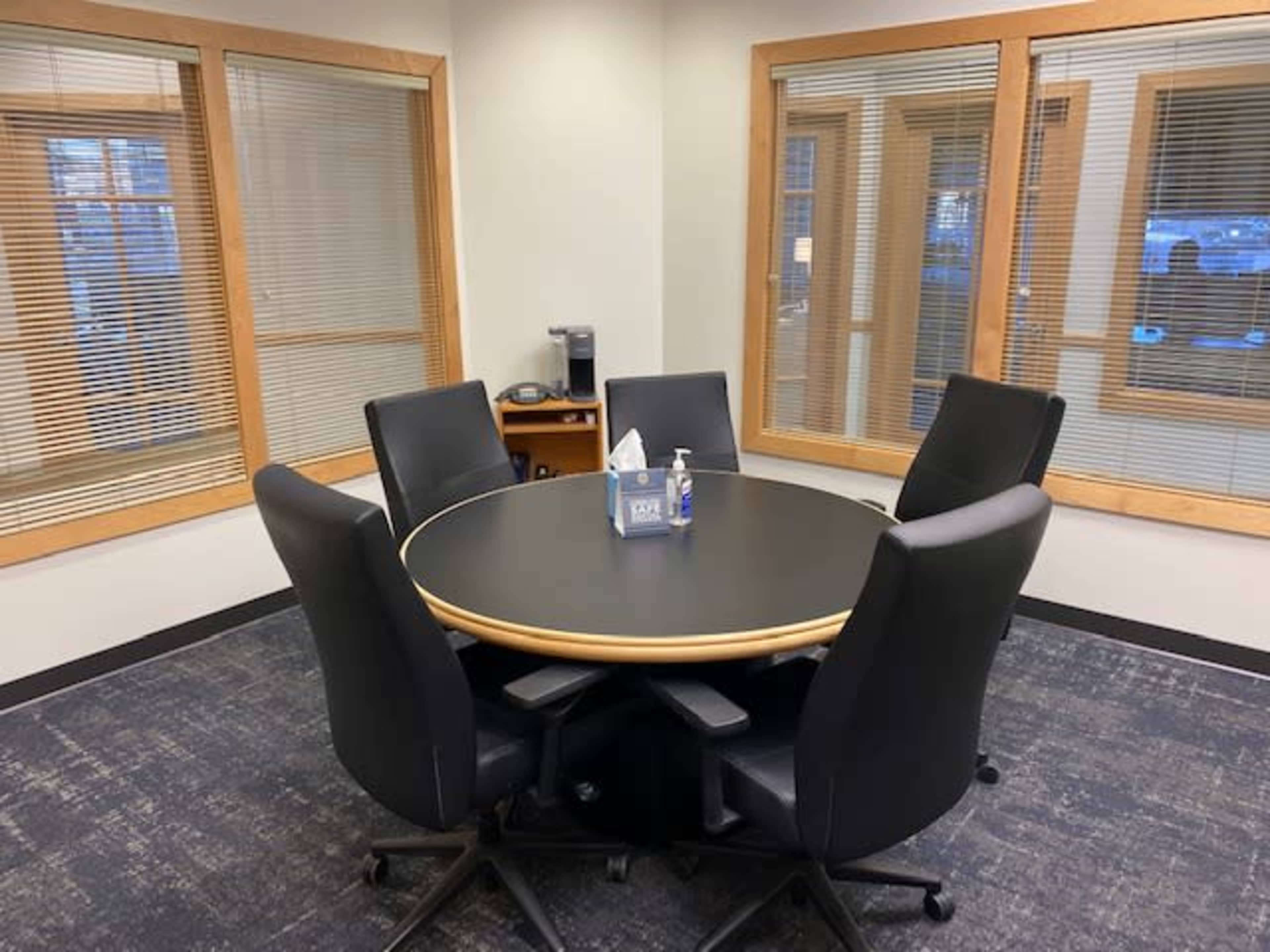 A round black conference table surrounded by five black office chairs in a well-lit meeting room with large windows.