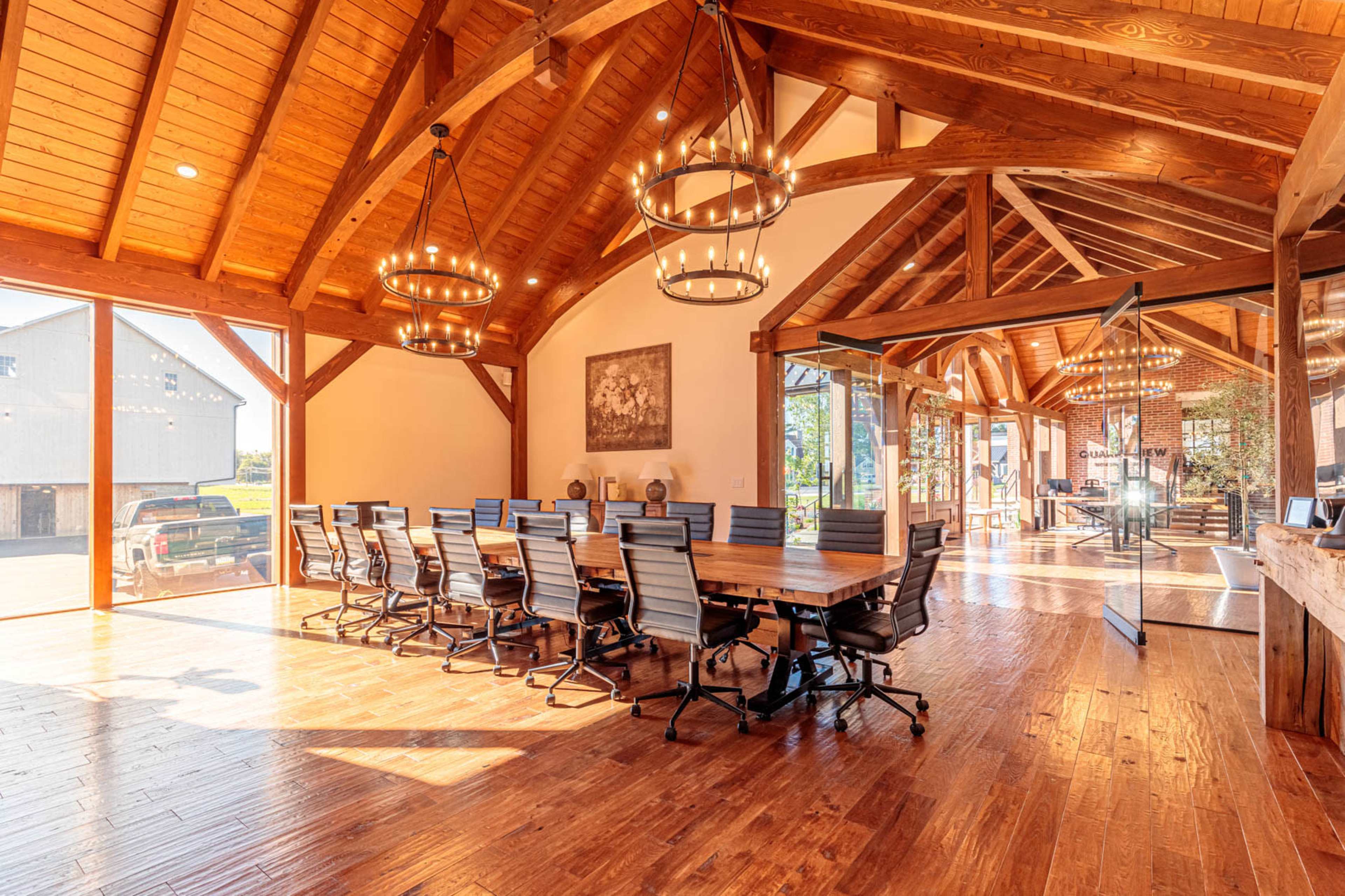 A spacious conference room features a large wooden table surrounded by black ergonomic chairs, with large windows showcasing an outdoor view and wooden beams overhead.