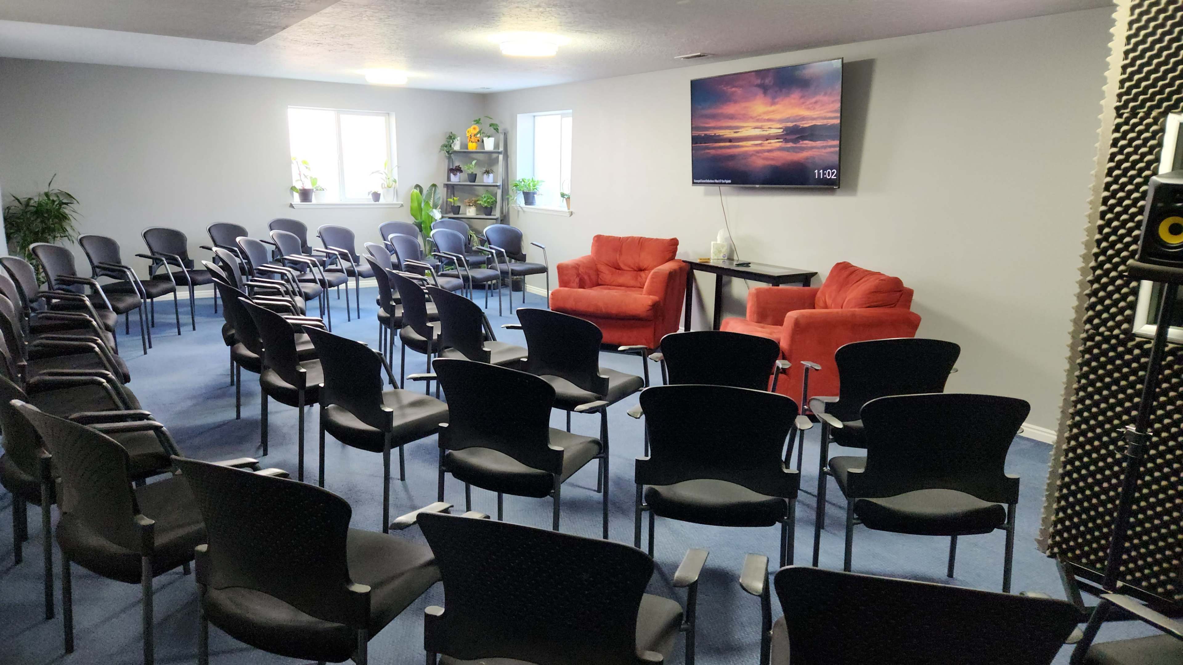The image shows a spacious room set up with rows of black chairs facing two red armchairs and a television mounted on the wall.