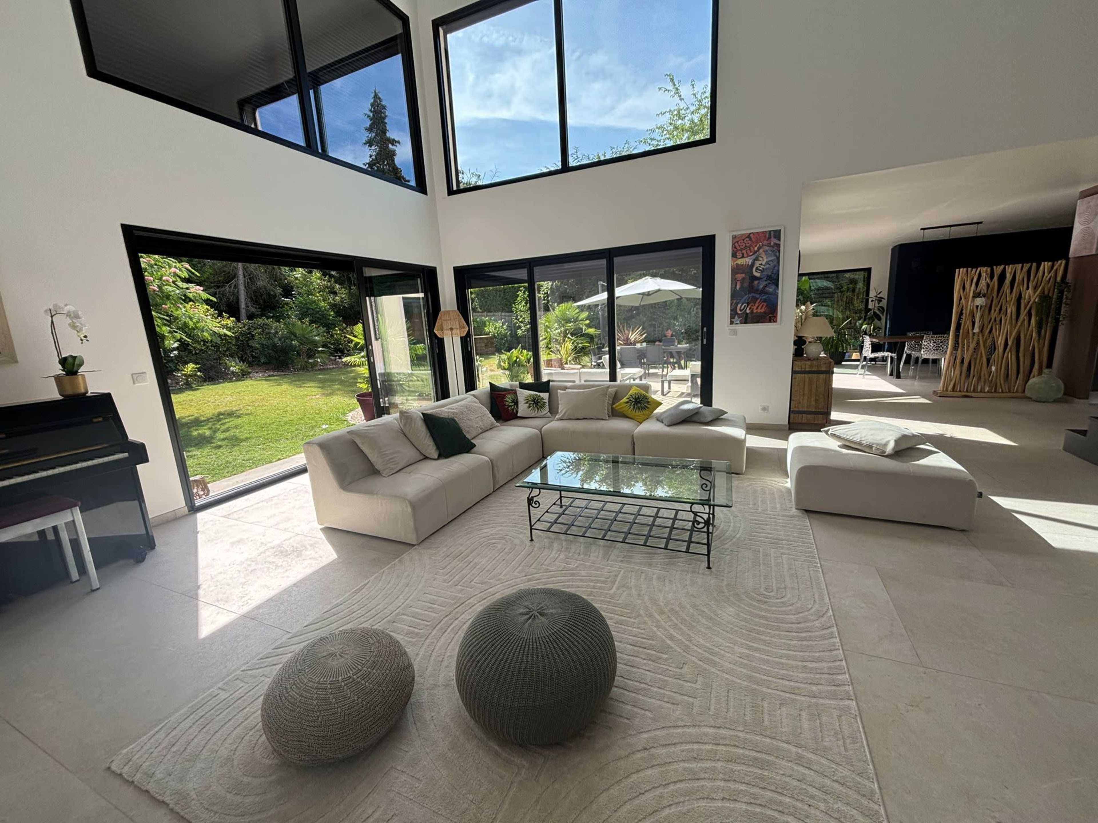 The image shows a modern living room with large windows overlooking a garden, featuring a sectional sofa, a glass coffee table, and two decorative poufs.