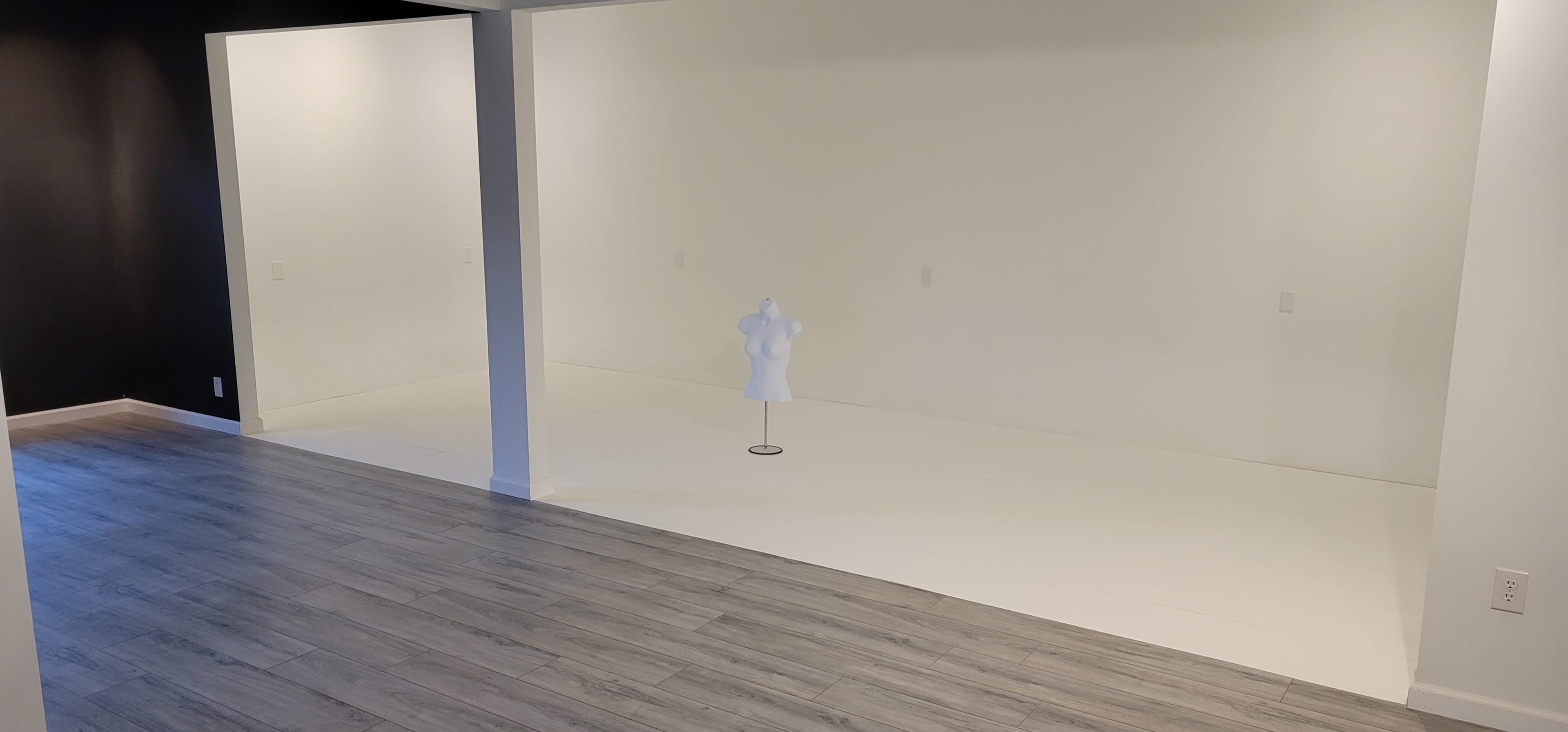 A spacious, empty room with white walls and a light wood floor, featuring a dress form positioned in the center.