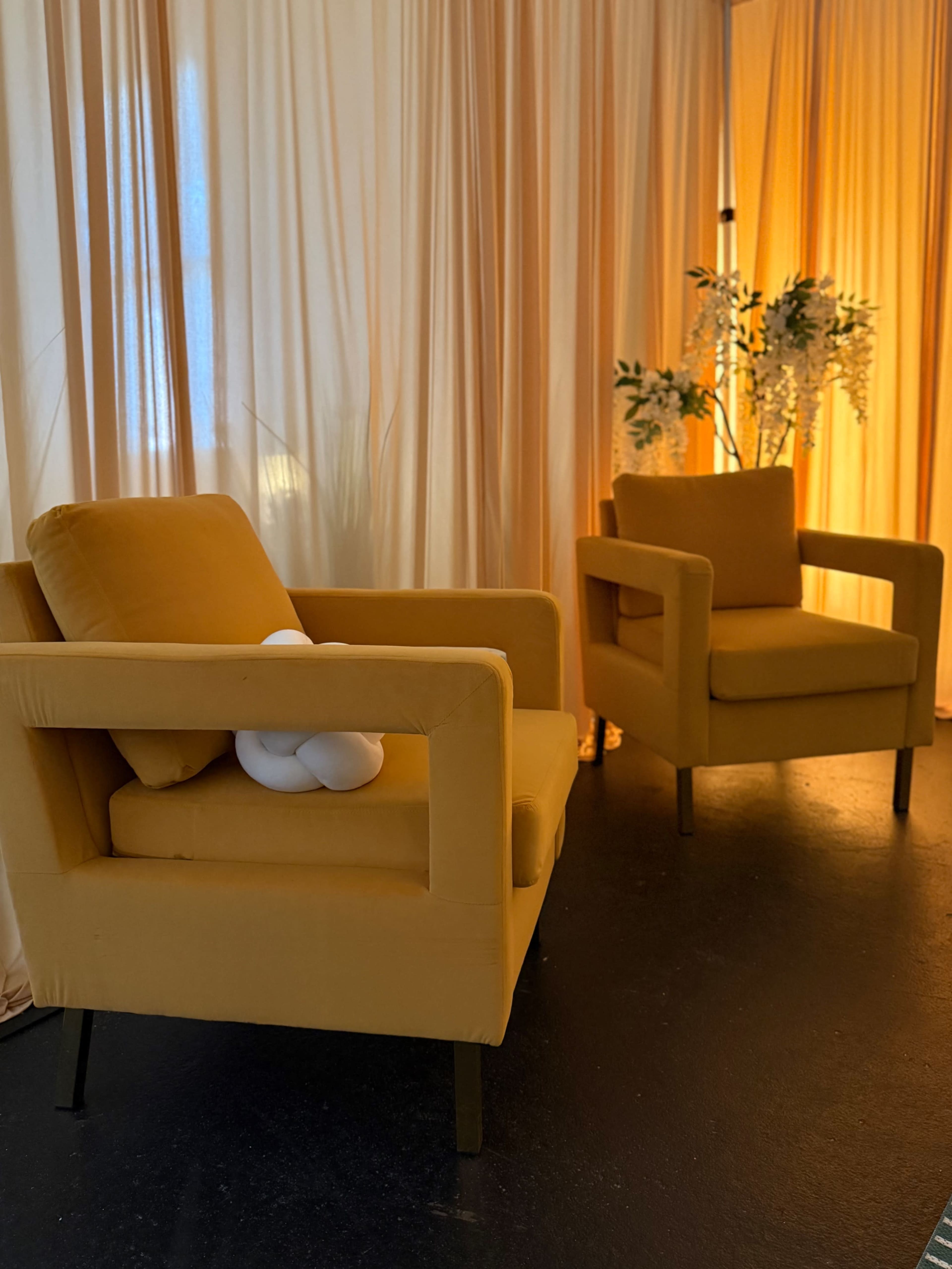 The image shows two mustard-colored armchairs placed against a backdrop of soft, flowing drapes with warm lighting.