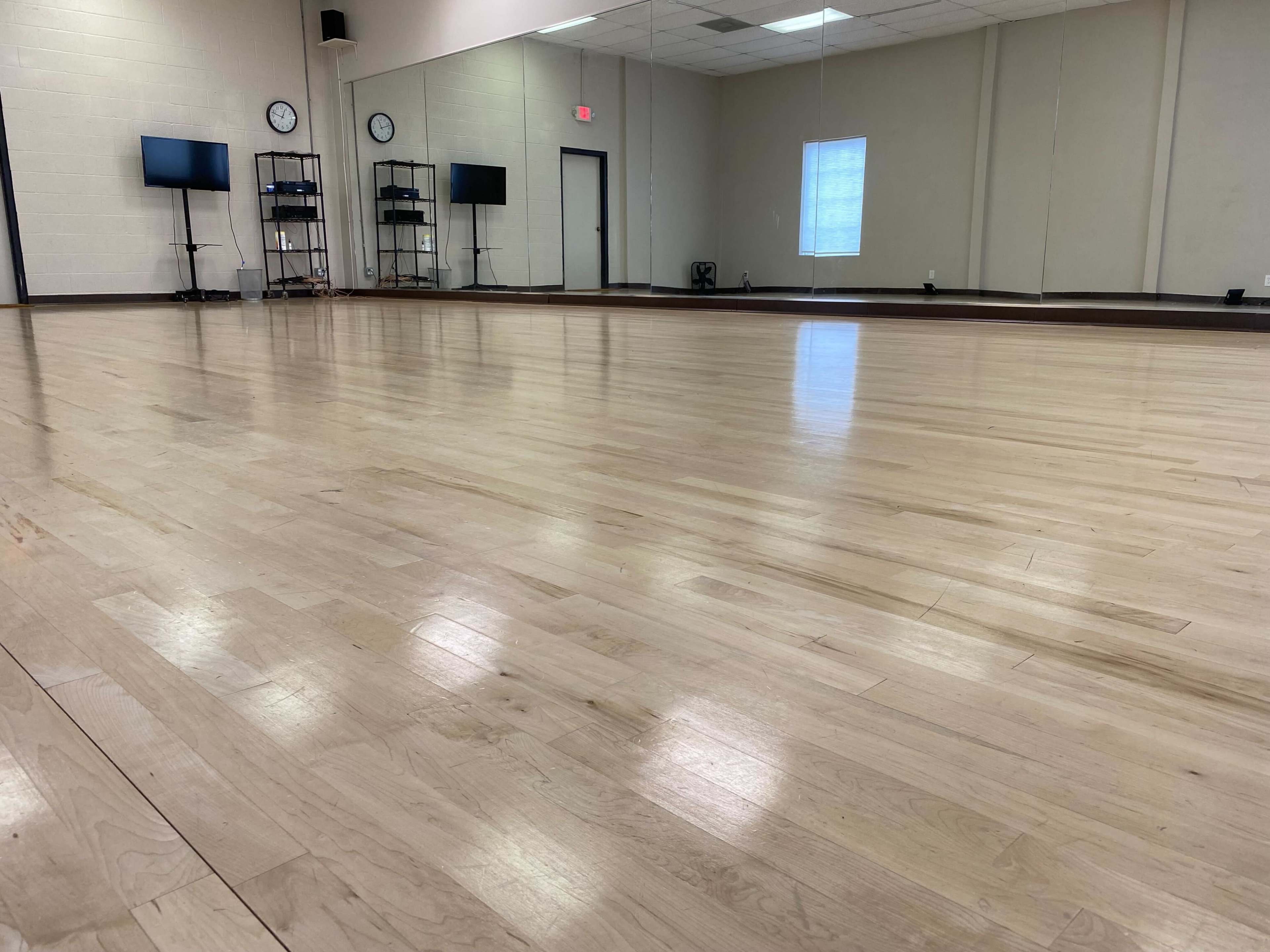 A spacious, empty dance studio with a polished wooden floor, large mirrors, and two wall-mounted clocks.