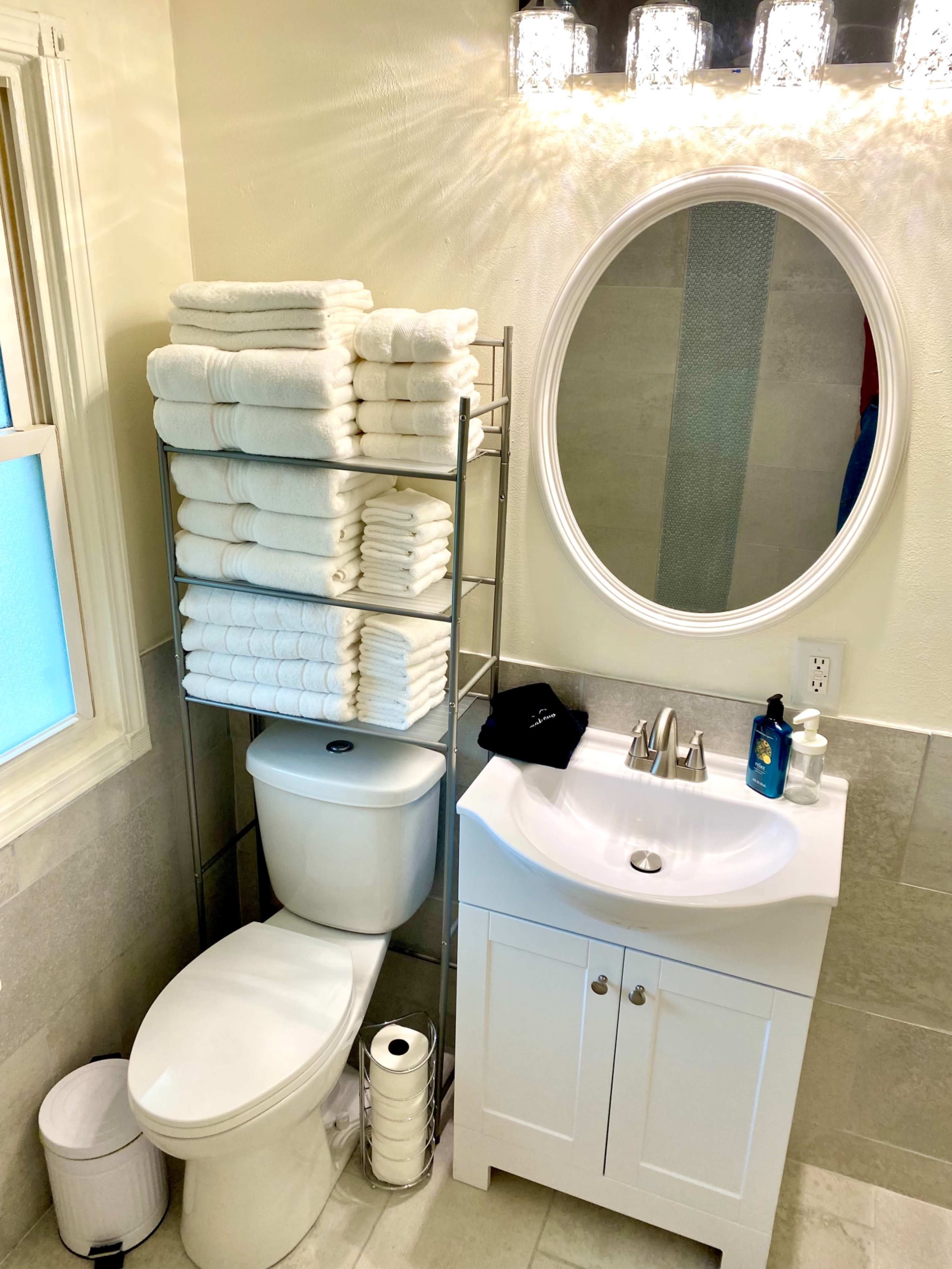 A neatly organized bathroom features stacks of white towels on a metal rack beside a toilet and a sink with a round mirror above it.