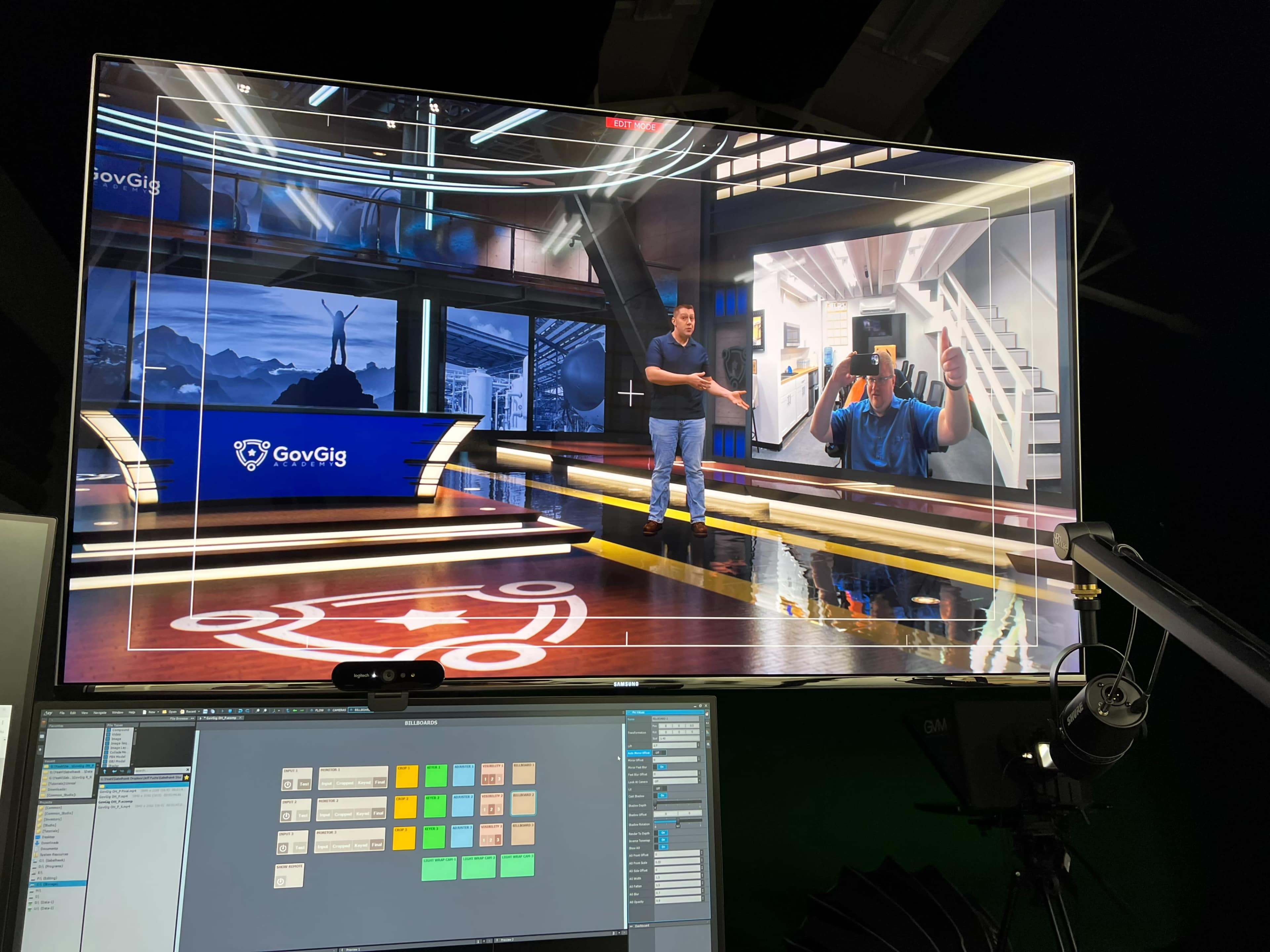 A person stands in a studio setting with a large monitor displaying another individual and graphics related to a presentation.