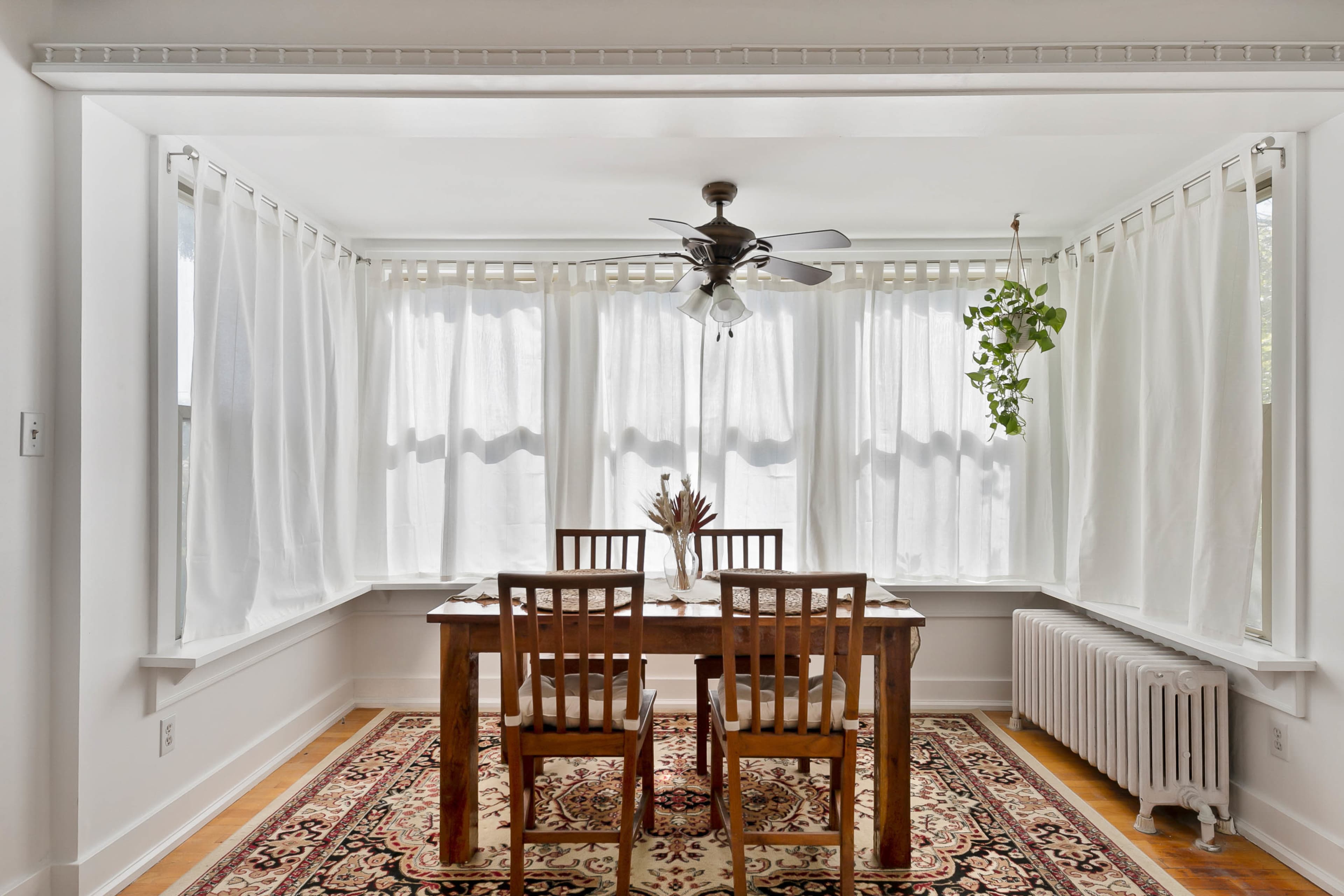 A dining area features a wooden table with four chairs, surrounded by large windows covered with sheer white curtains and a radiator against the wall.