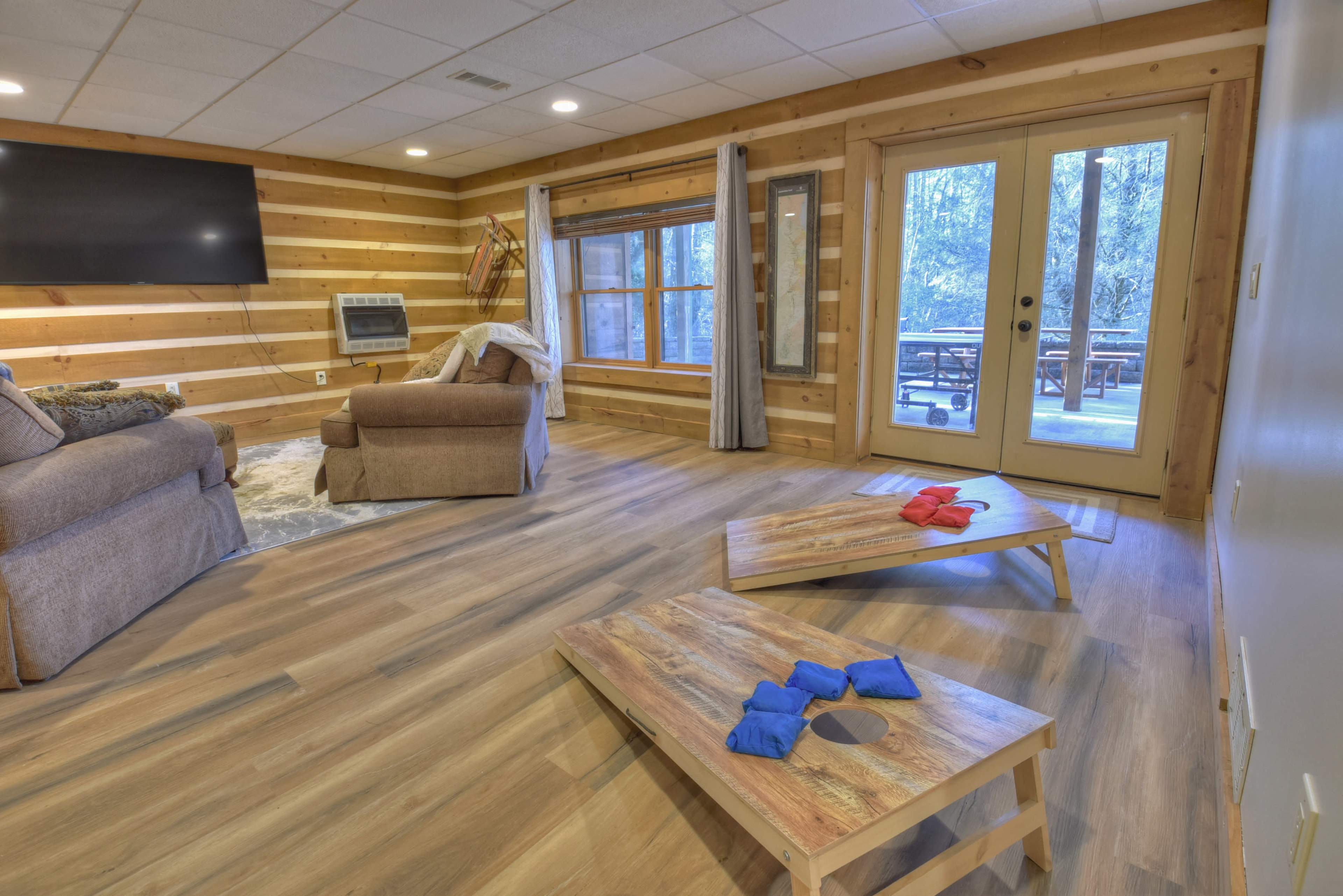 The room features wooden walls, a large TV, a cozy seating area with a recliner and sofa, and a set of colorful game boards on the floor near a set of glass doors leading to an outdoor deck.