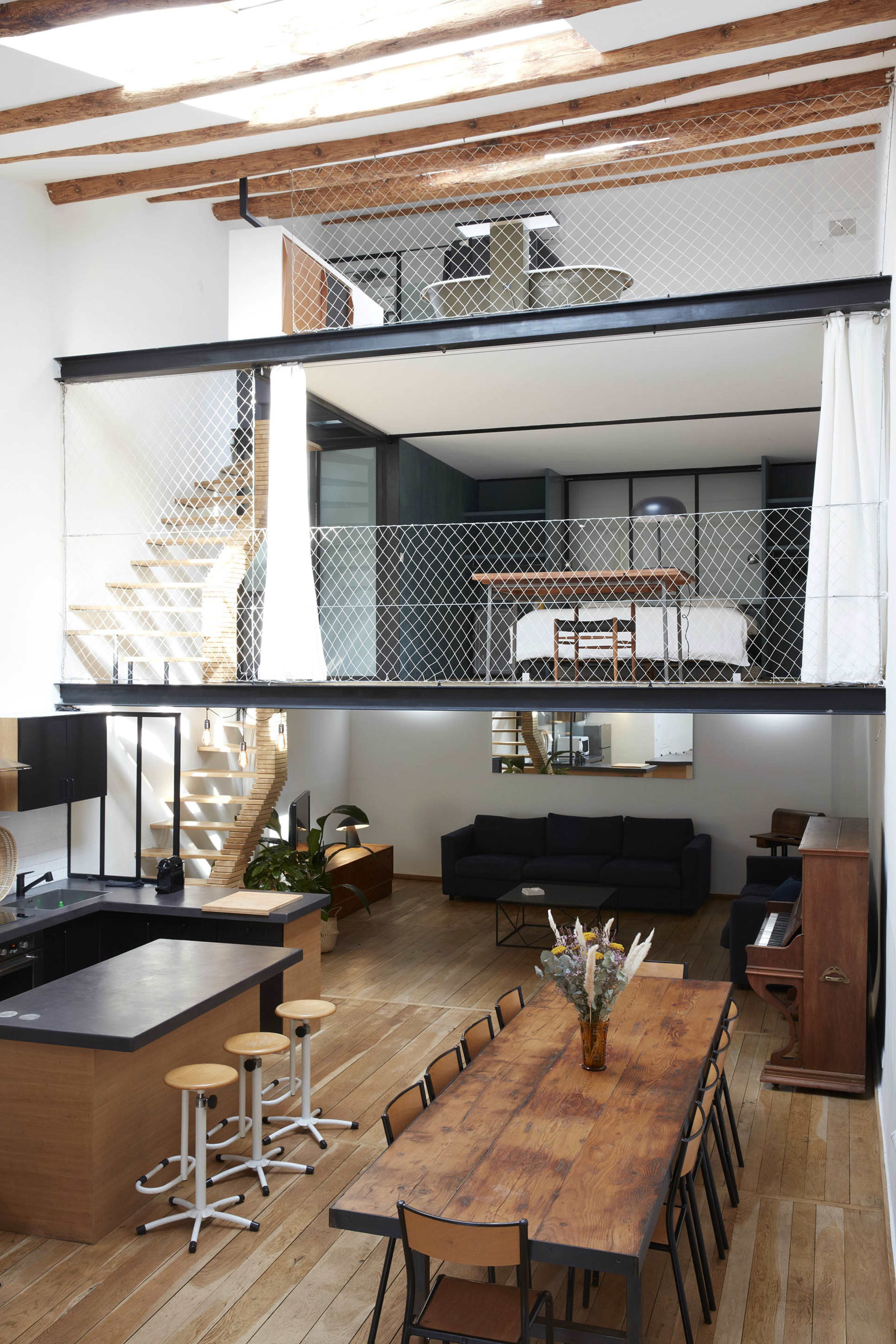 The image features a modern, multi-level interior of a house with an open-plan design that includes a kitchen, dining area, and a living space, accentuated by a staircase and a lofted bedroom above.