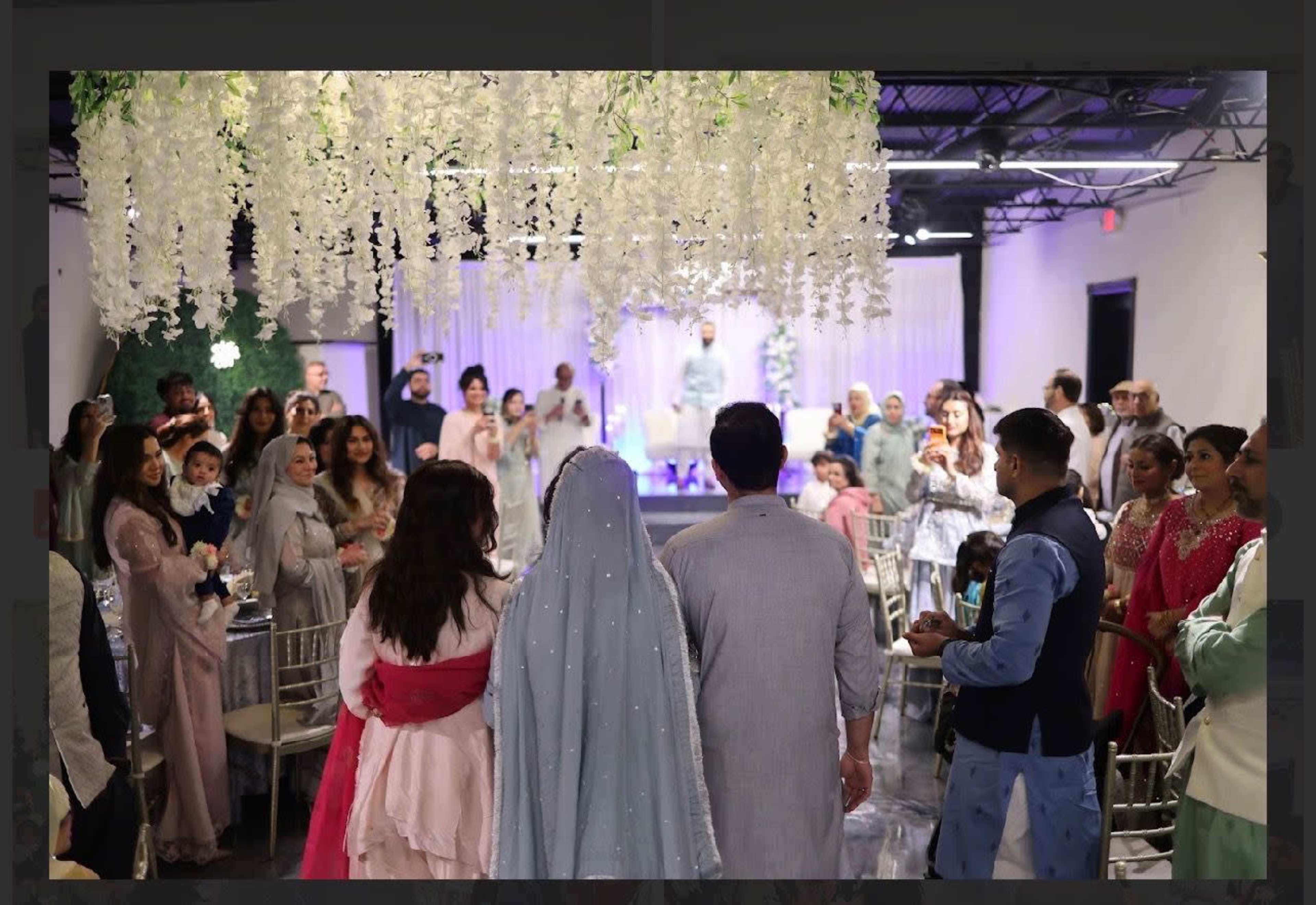 A group of elegantly dressed people gather in a decorated venue with floral arrangements for a celebration.