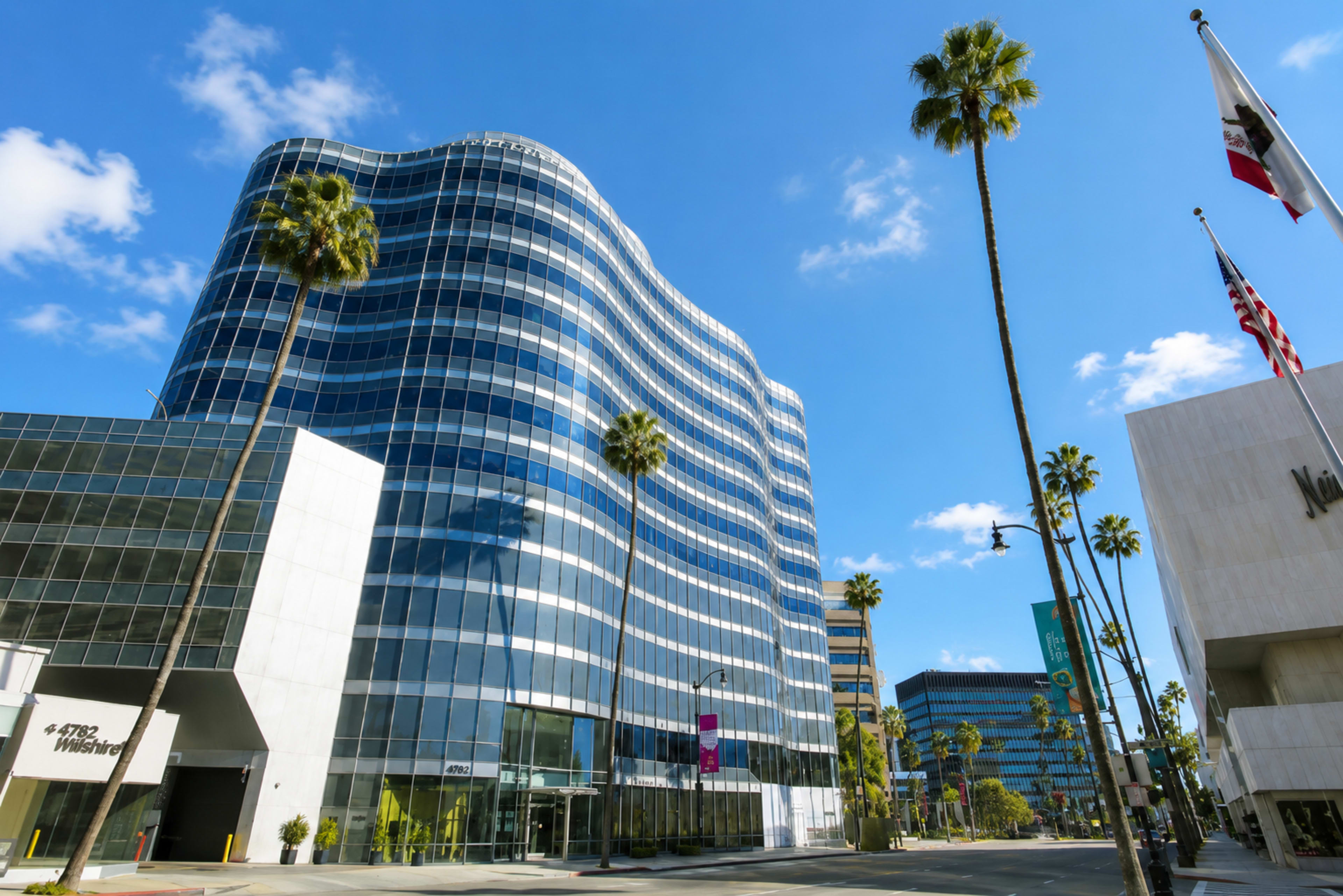 The image shows a modern glass building with a wavy design, surrounded by palm trees and other structures, under a clear blue sky.