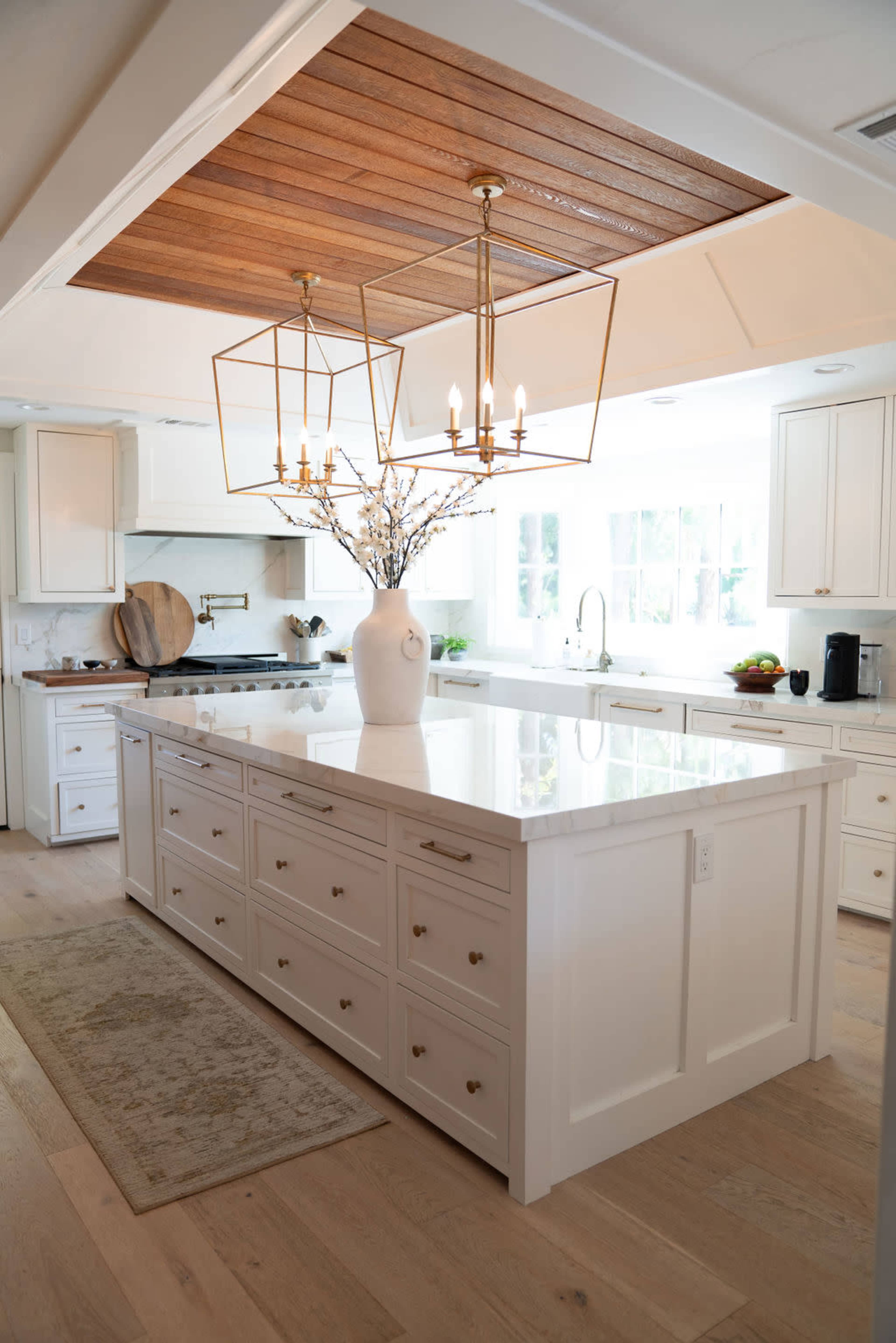 A modern kitchen features an island with multiple drawers, a large vase with branches as a centerpiece, and pendant lighting above a wooden ceiling.