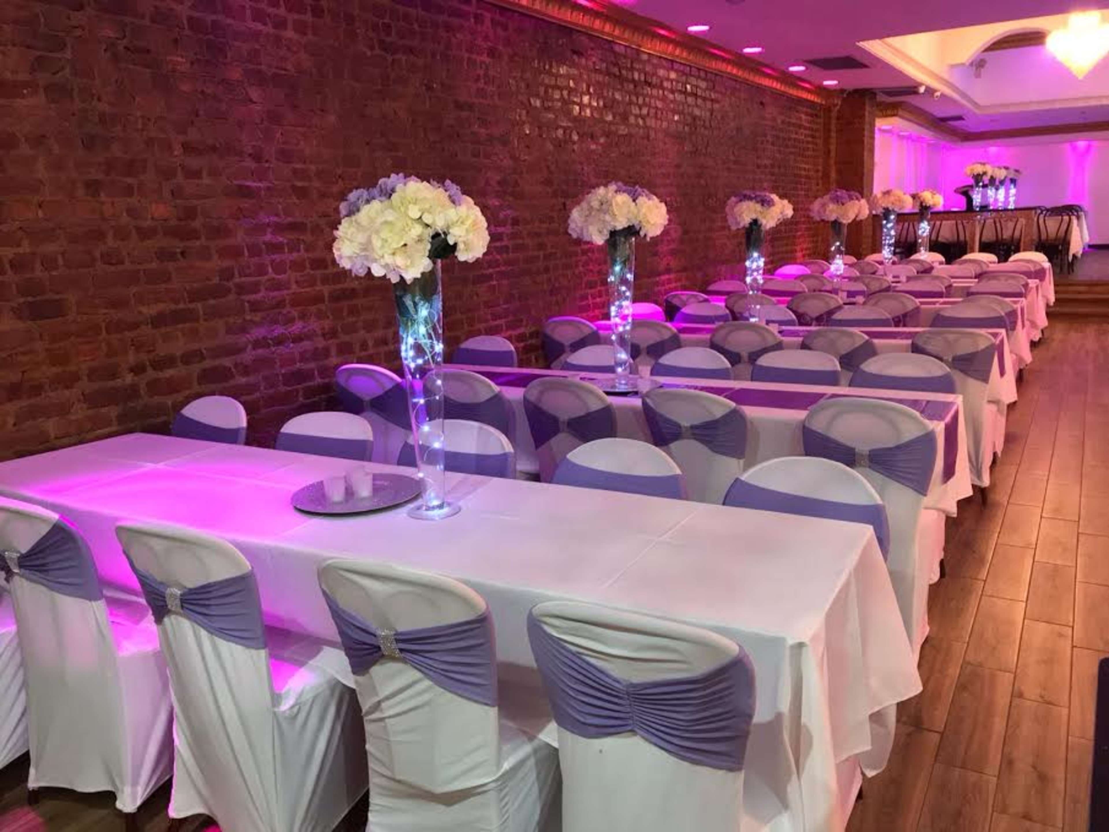 A banquet hall features long tables covered in white cloths with purple sashes, adorned with floral centerpieces and illuminated by soft lighting.