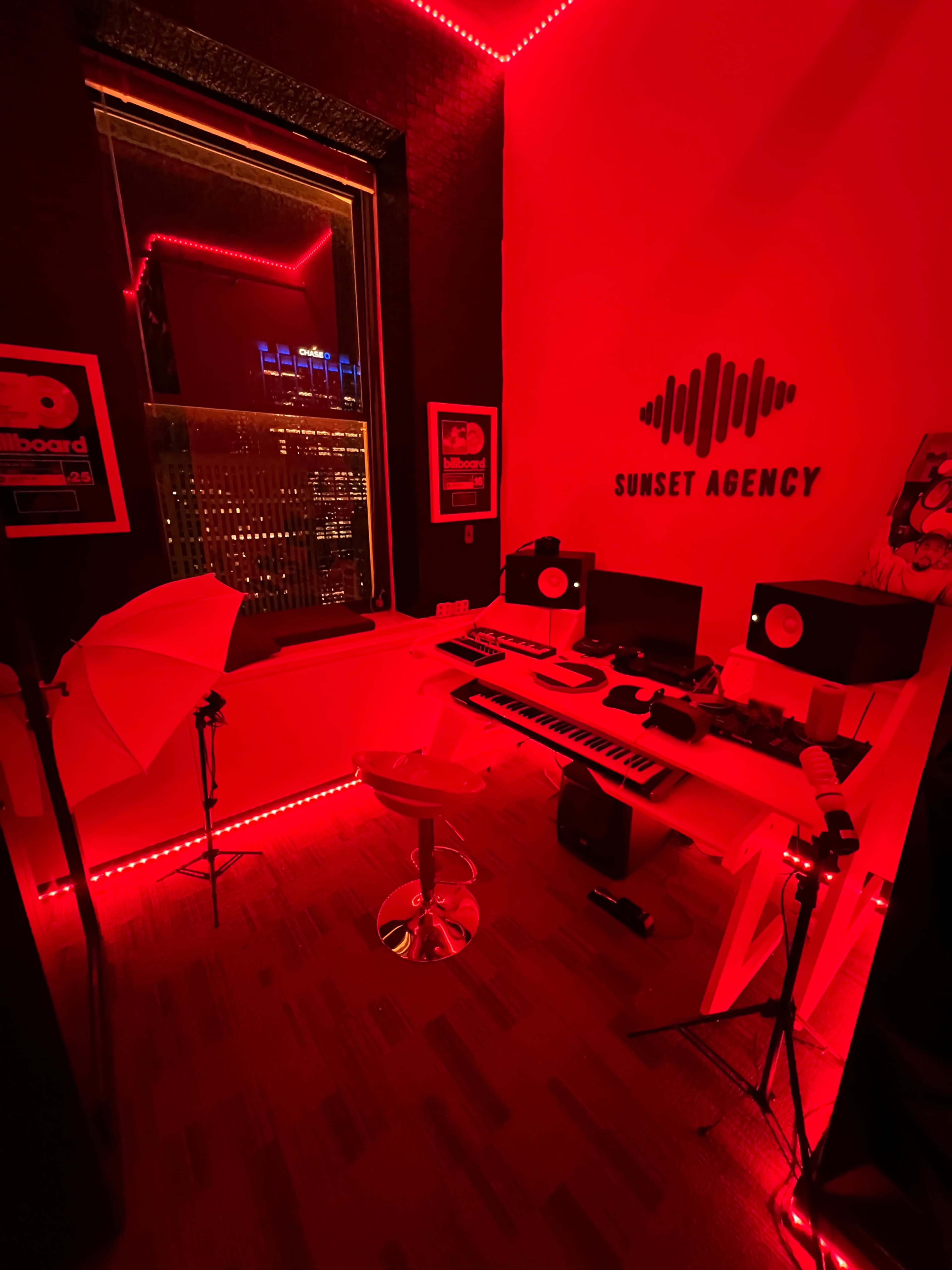 A music production studio features a white desk with a keyboard, speakers, and recording equipment, illuminated by red LED lights.