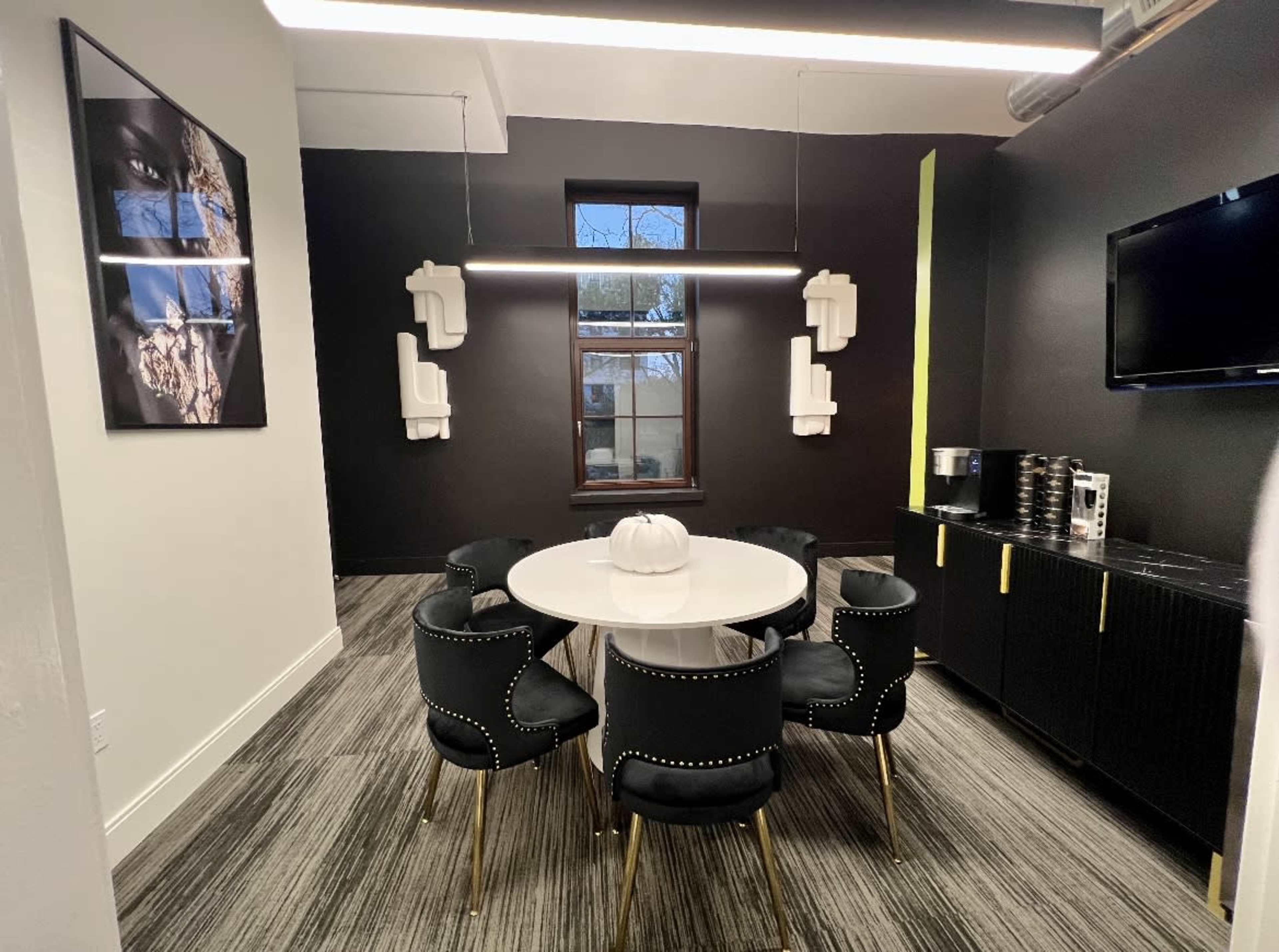 A modern meeting room features a circular white table surrounded by six black chairs with gold accents, along with a wall-mounted television and artwork.