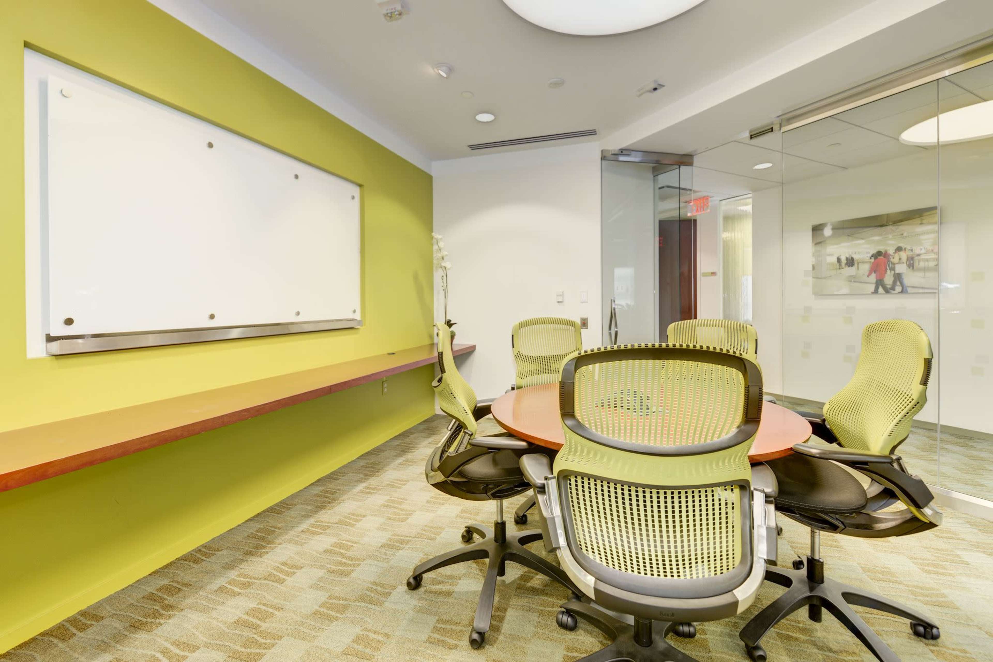 A conference room features a round table surrounded by several ergonomic chairs and a whiteboard against a vibrant green wall.