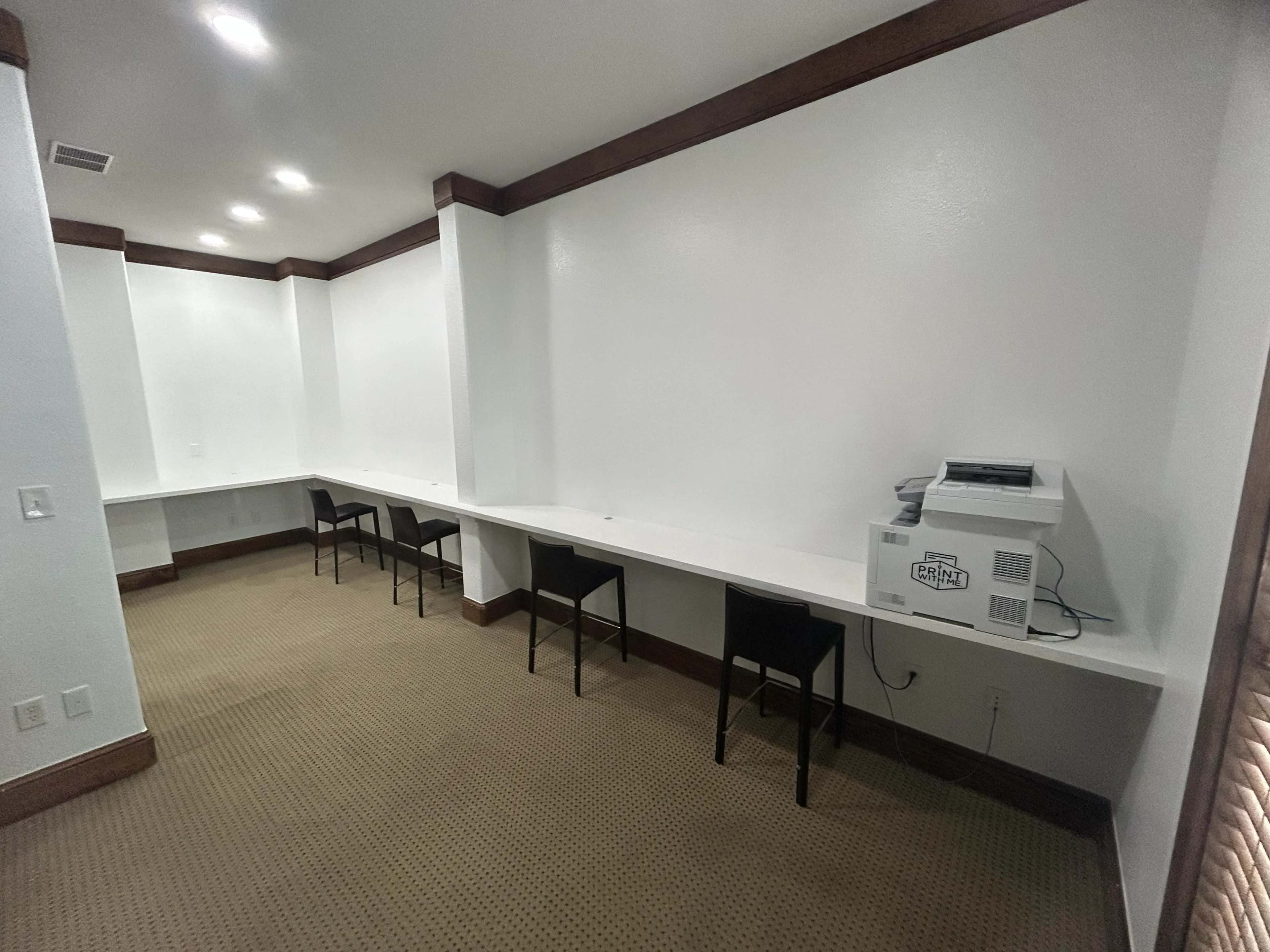 The image shows a sparsely furnished office space with a long white countertop and several black chairs, accompanied by a printer in the corner.