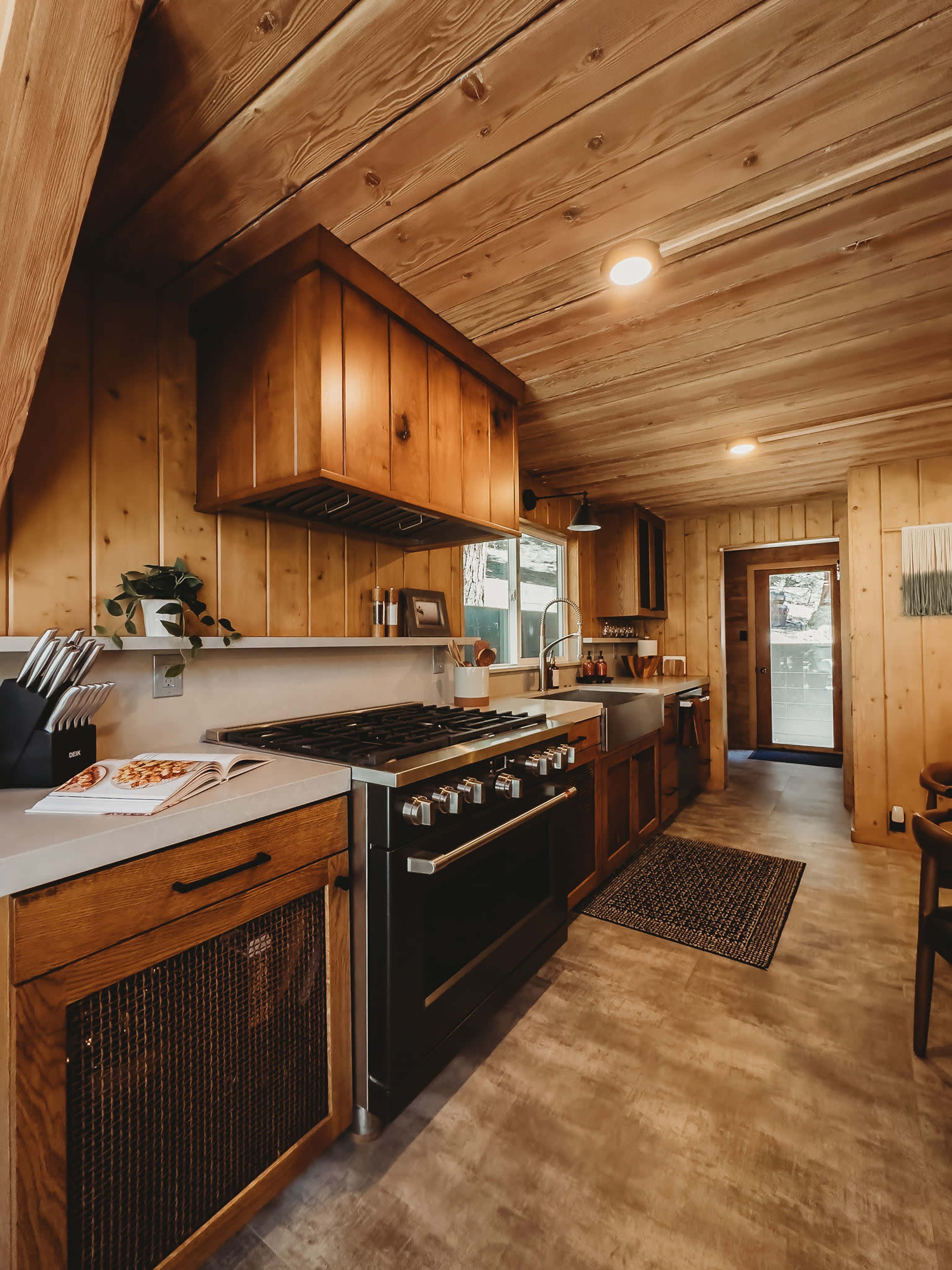 The image depicts a modern kitchen with wooden cabinetry, a large gas stove, and a view of a door leading outside.