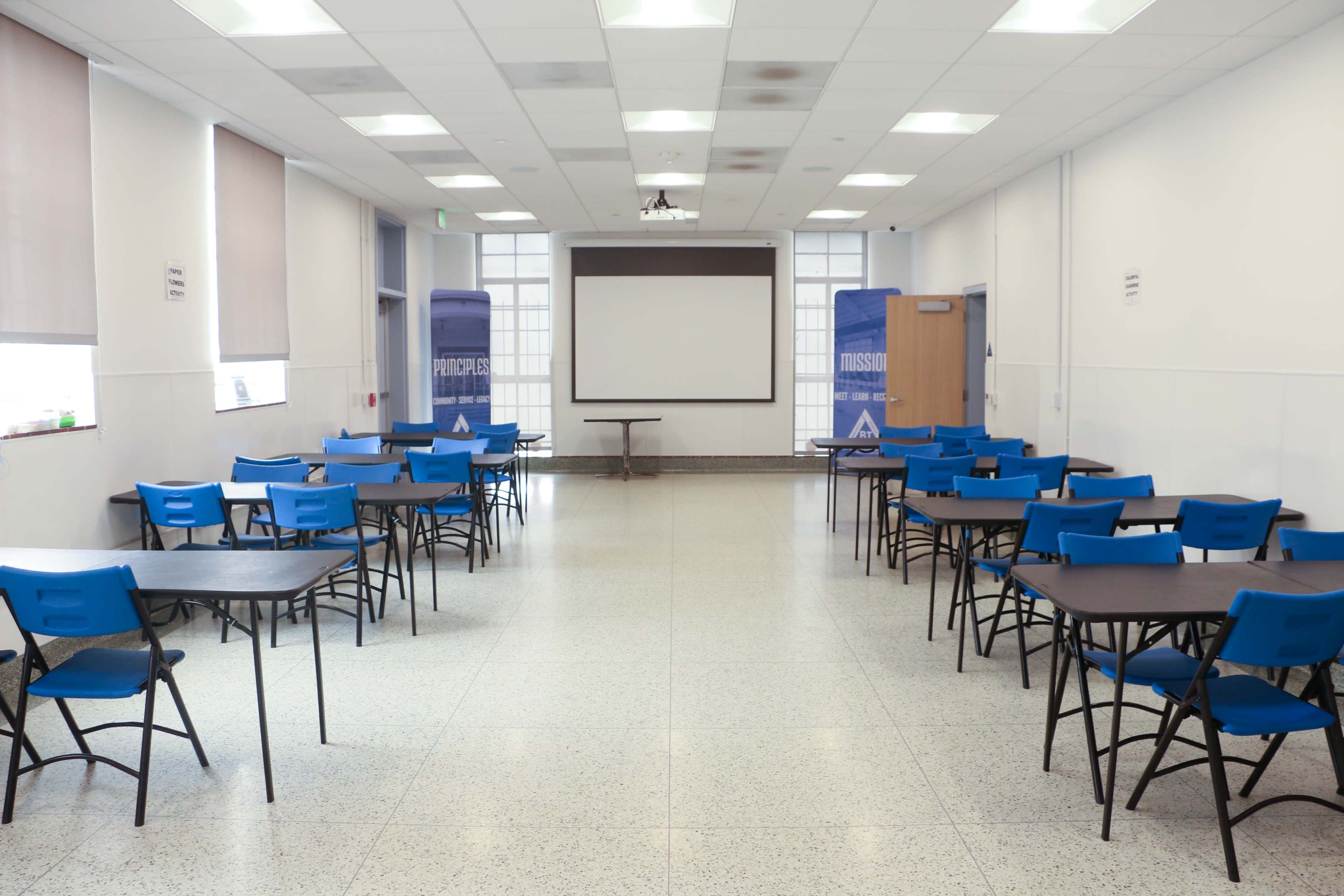 A spacious classroom is set up with multiple blue chairs and tables arranged for group activities, featuring a projector screen at the front.
