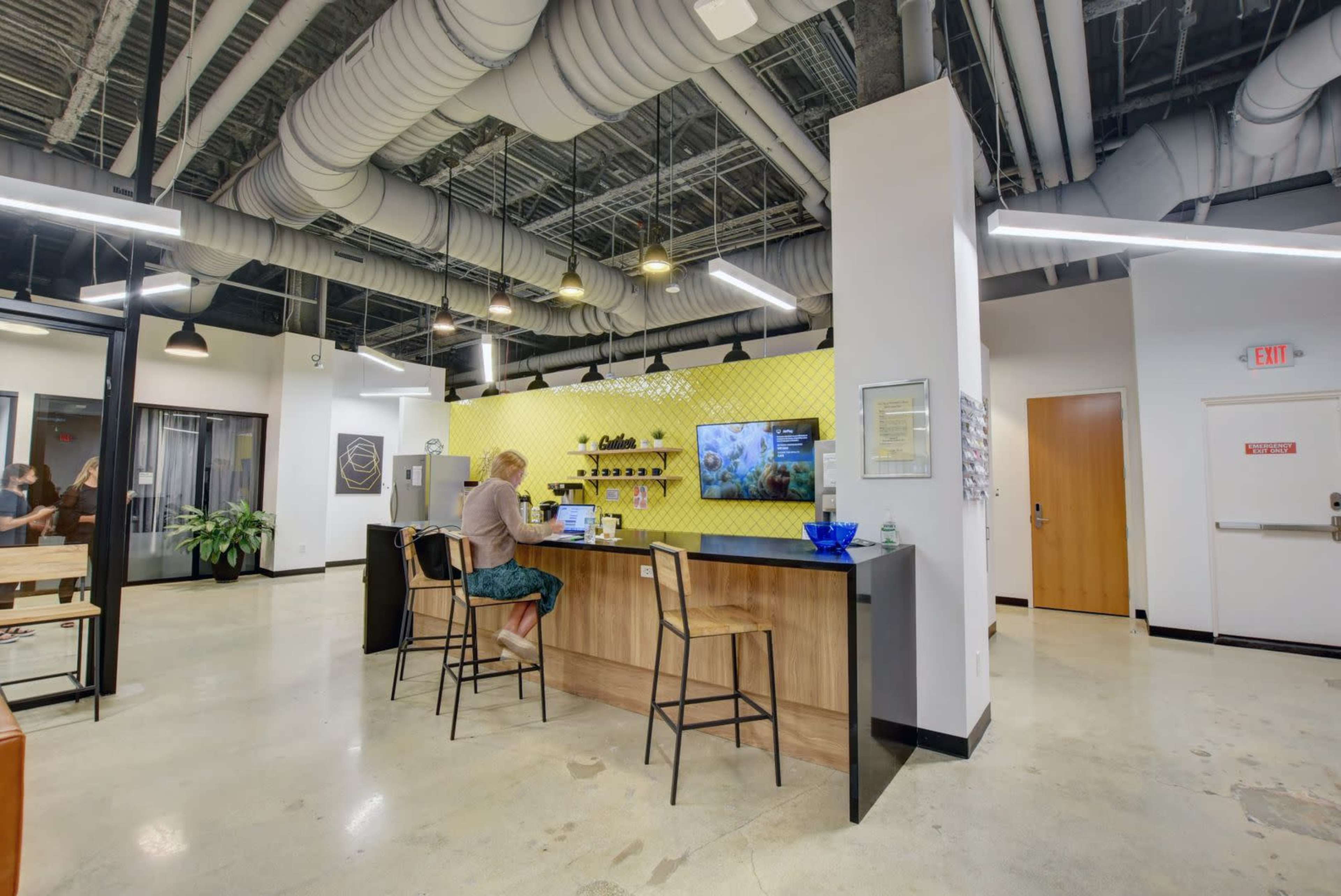 A person is working at a counter in a modern office space with yellow and gray decor, featuring industrial-style ceilings and plants.