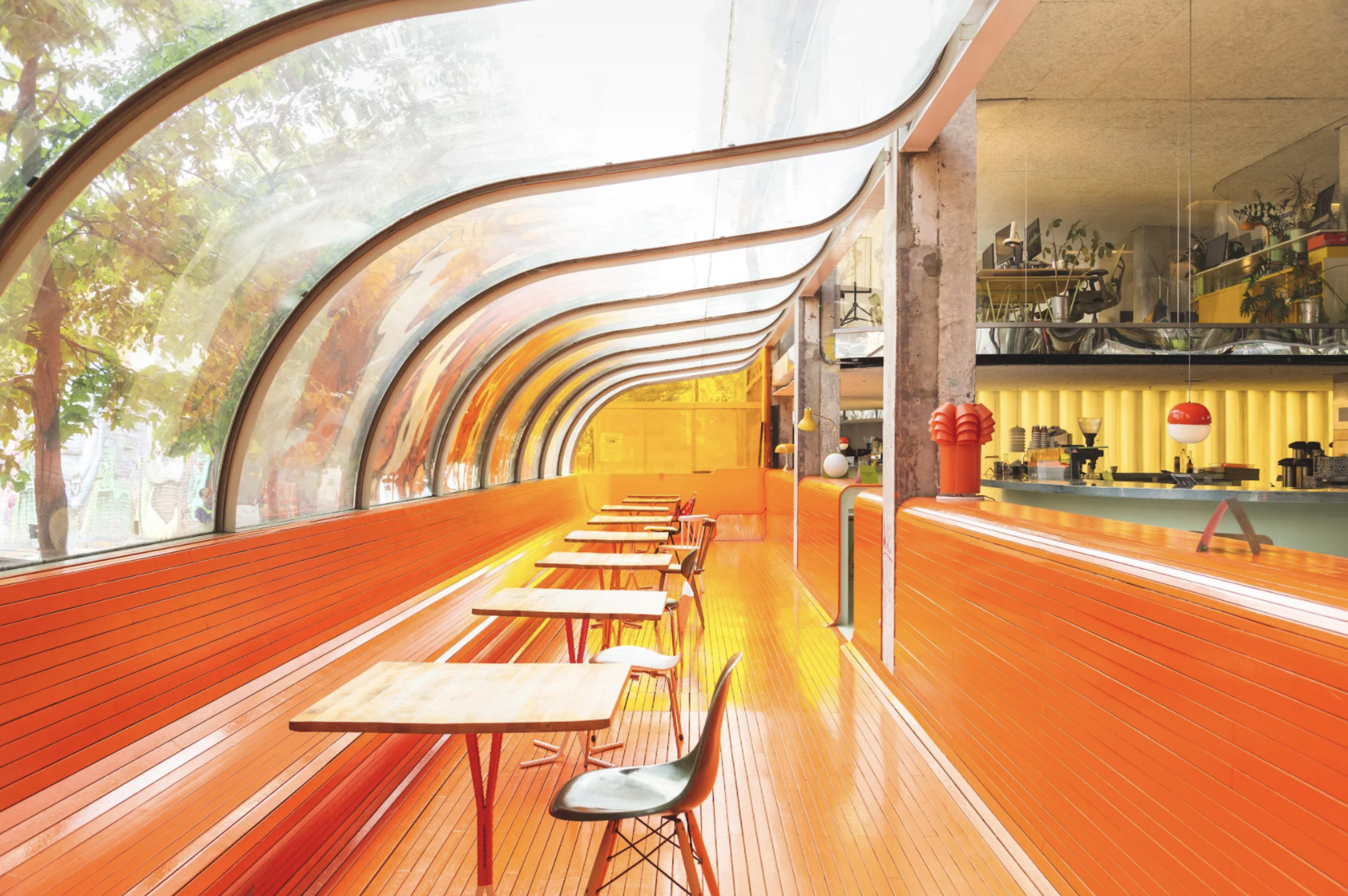 A modern indoor café with curved glass walls and an orange wooden interior, featuring empty tables arranged along the space.