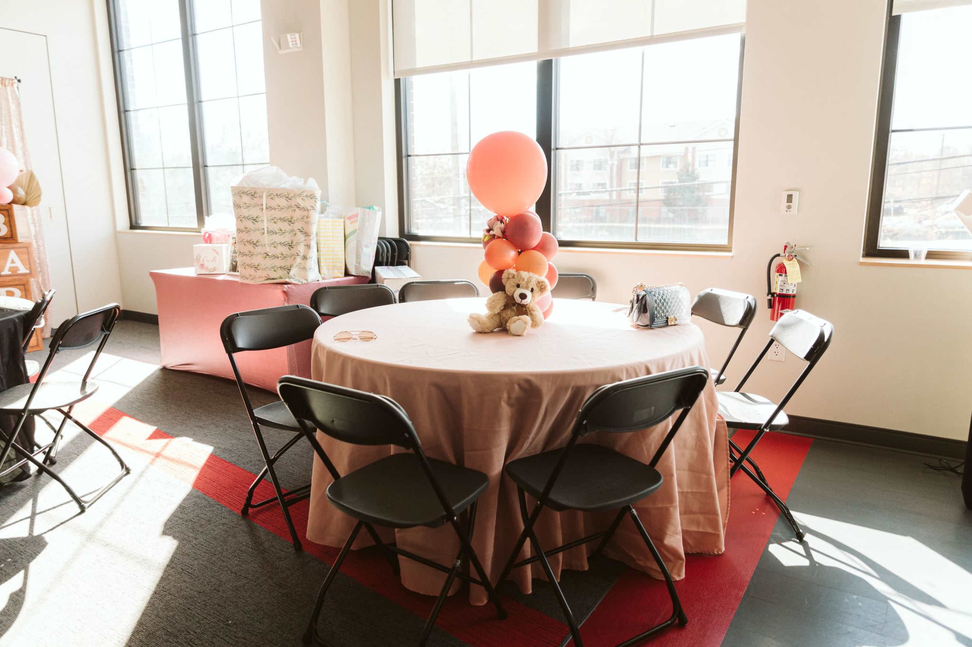 A round table with a beige cloth is set in a brightly lit room, surrounded by black chairs, and features a teddy bear and a cluster of balloons.