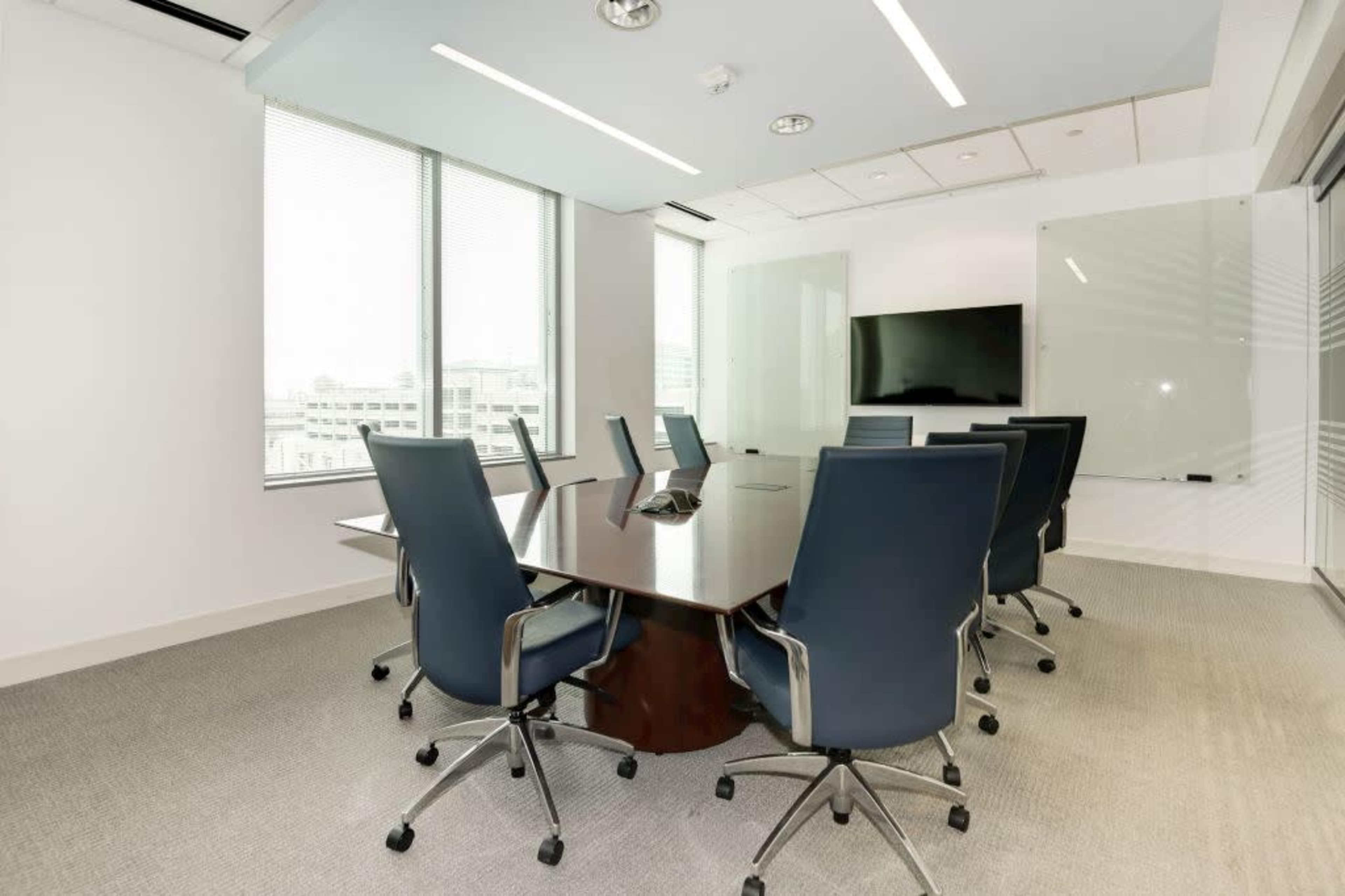 A modern conference room features a large wooden table surrounded by eight blue ergonomic chairs, with floor-to-ceiling windows and a wall-mounted television.