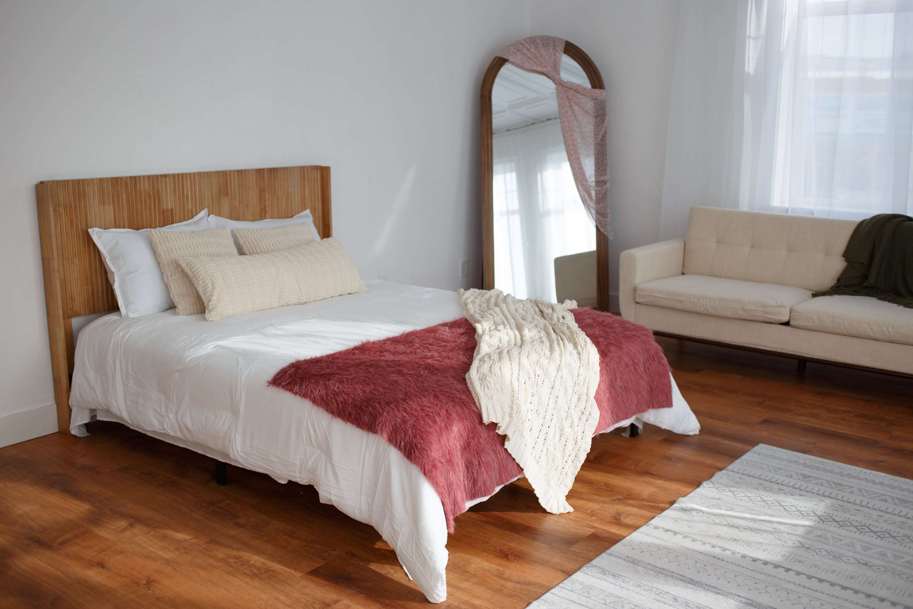 The image shows a well-lit bedroom featuring a bed with a wooden headboard, a cozy throw draped over the bed, a large mirror, and a minimalist sofa.