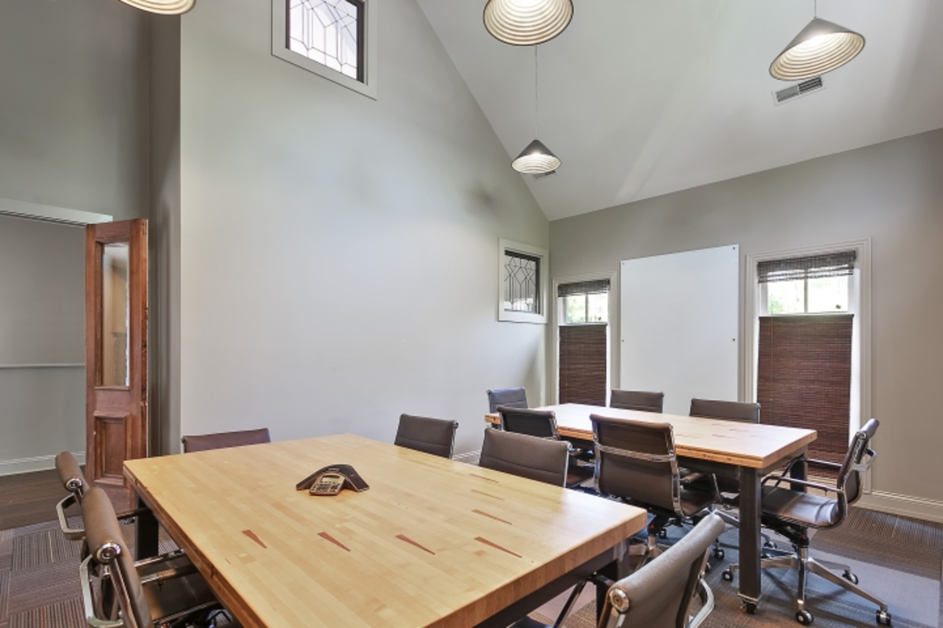 A conference room features a large wooden table surrounded by chairs, with windows and modern lighting fixtures.