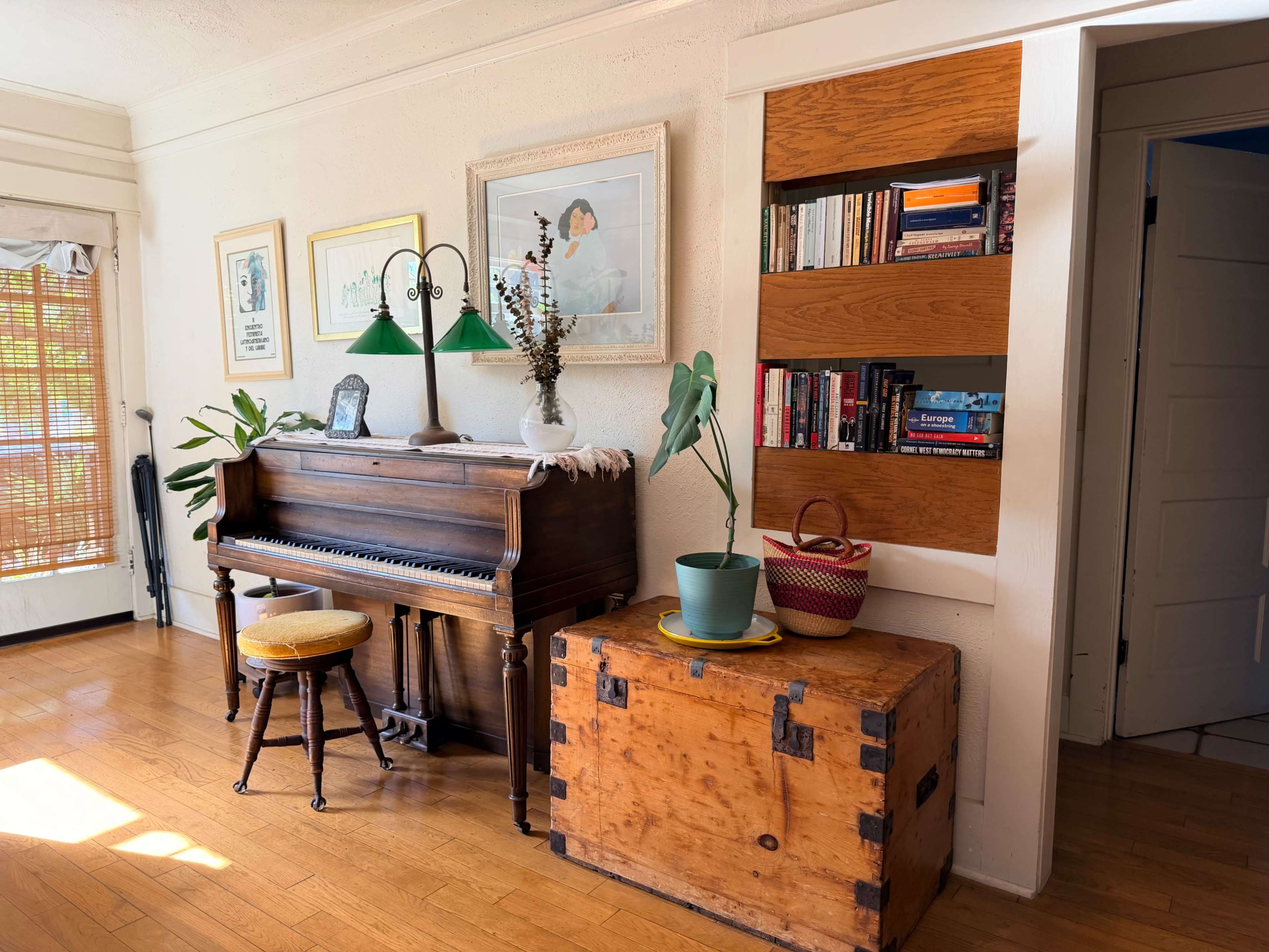The scene features a wooden piano with a stool beside it, complemented by a bookshelf and a large trunk against the wall in a well-lit room.