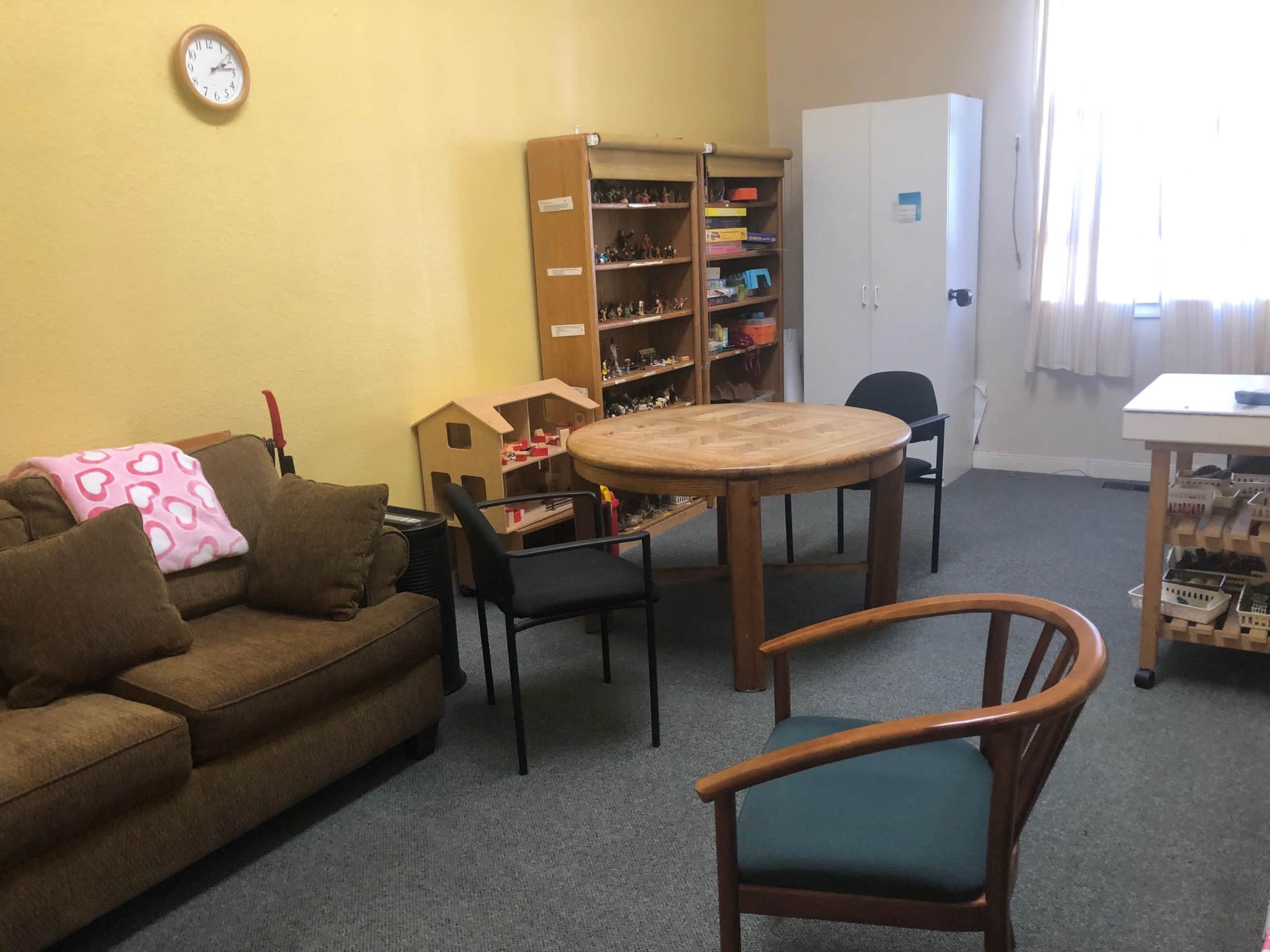 The image depicts a small room with a circular table surrounded by several chairs, a couch, a bookshelf filled with toys, and a white cabinet.