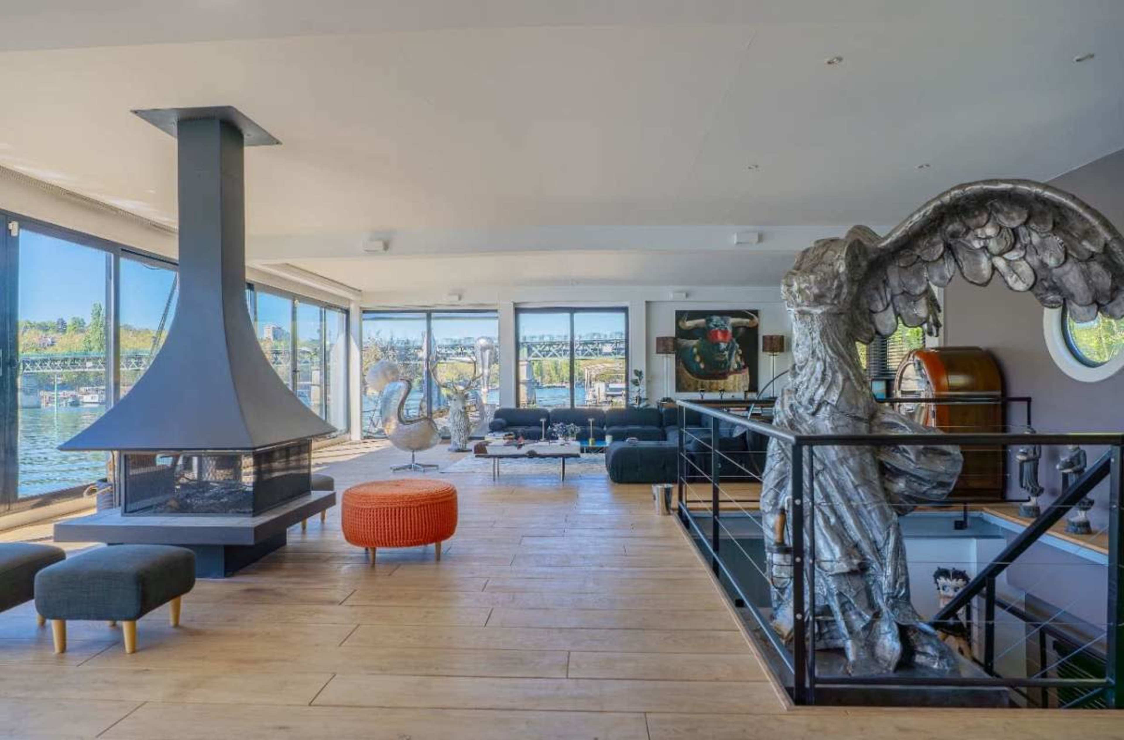 The image shows a contemporary living space with large windows, a central fireplace, a statue of an angel, and a lake view outside.