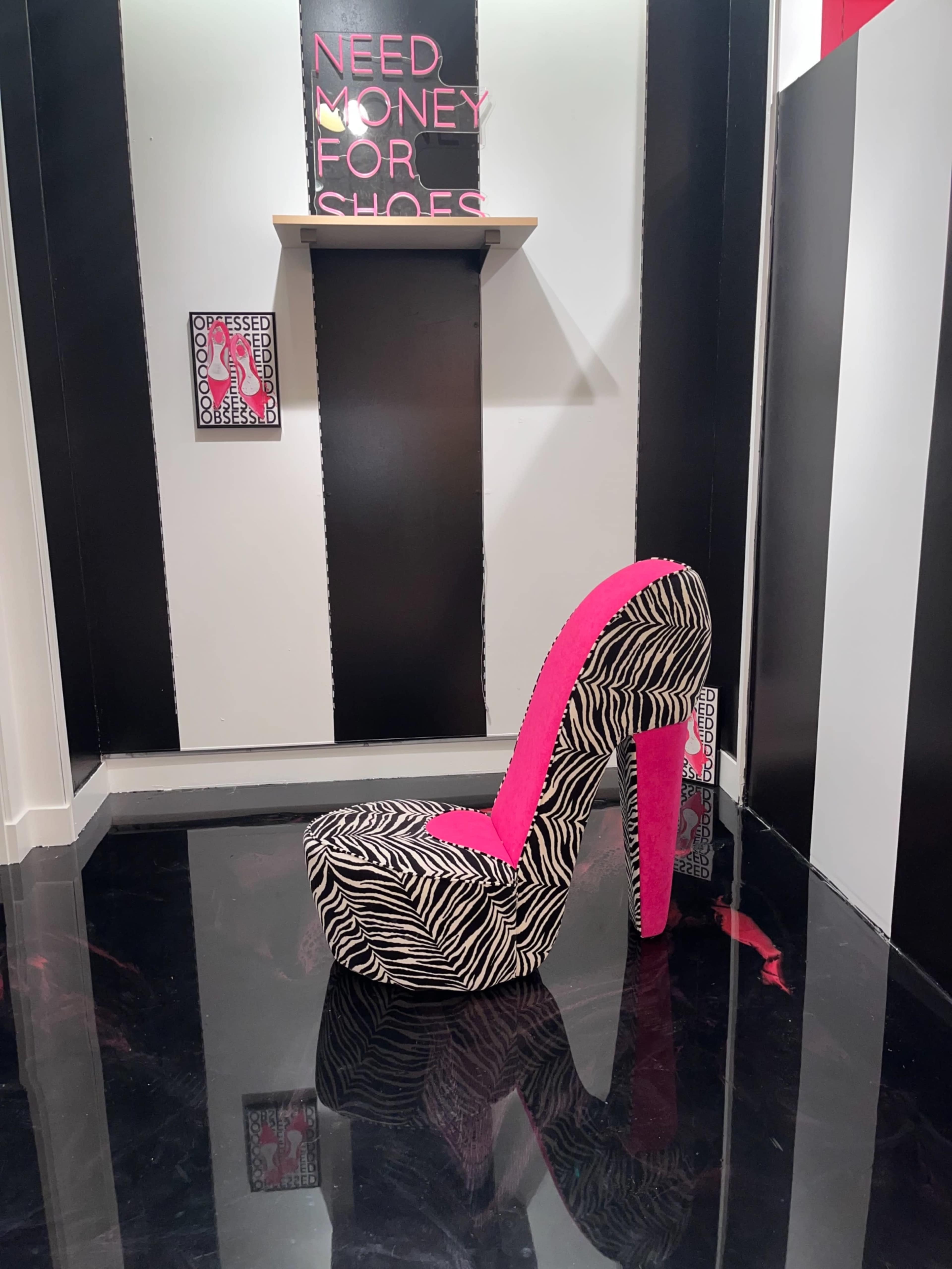 A large, zebra-striped high heel shoe with a pink lining is placed on a glossy black floor in a room adorned with bold black and white walls and a shelf displaying a sign that reads "NEED MONEY FOR SHOES."