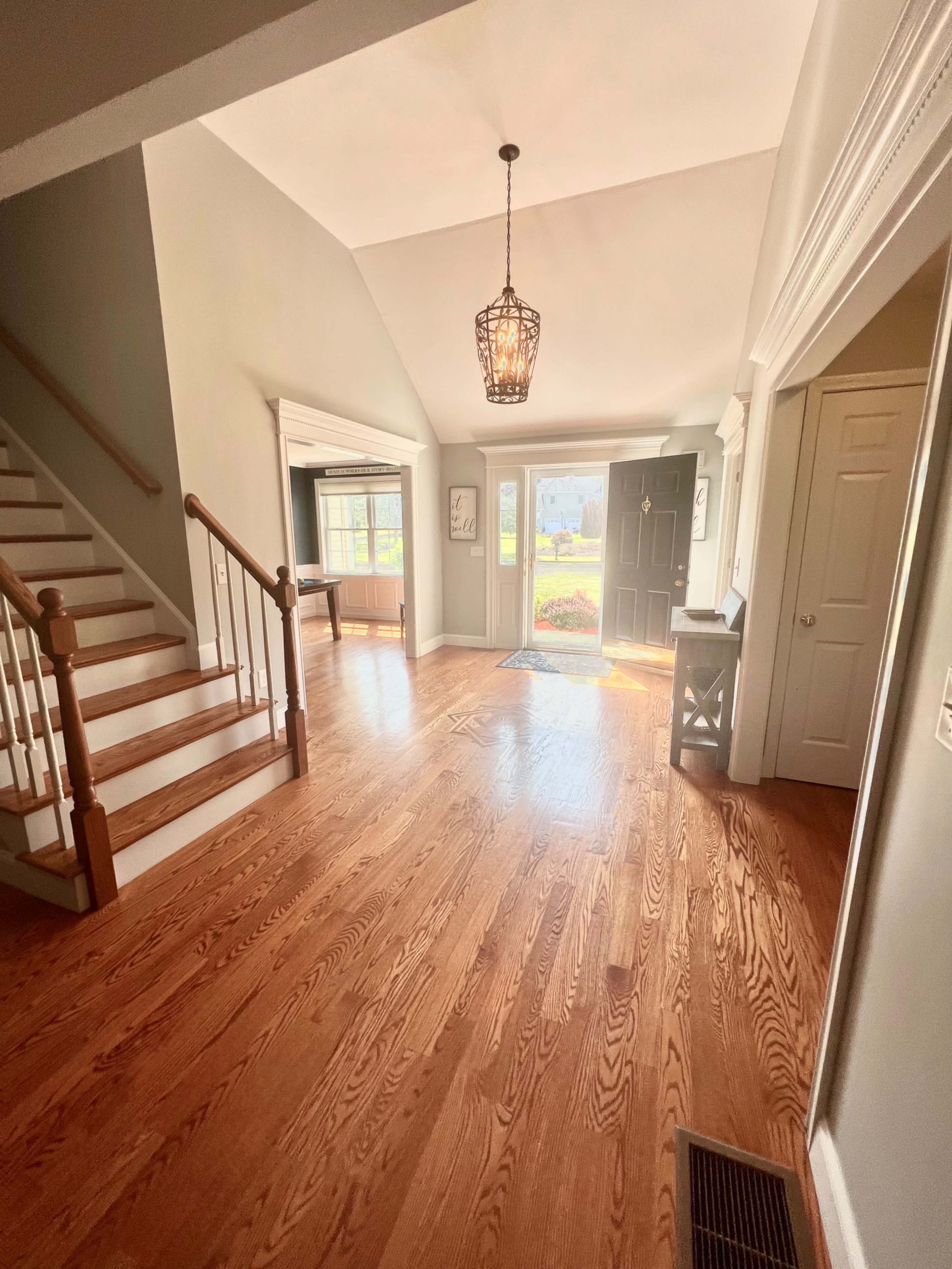A bright entryway with a staircase on the left, a front door at the center, and wooden flooring throughout.