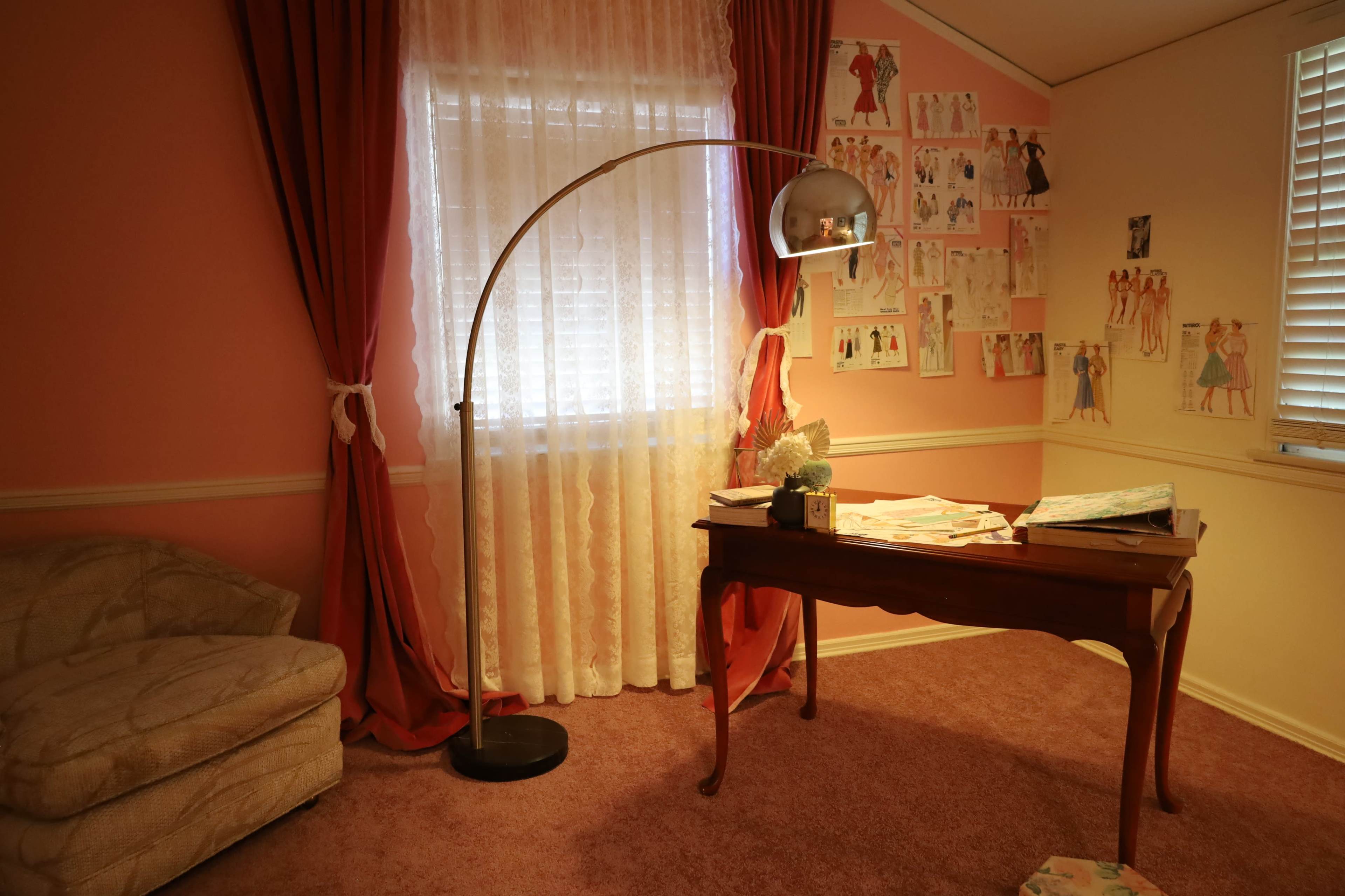 The room features a pink color scheme with a large window draped in sheer curtains, an arc floor lamp, a wooden desk covered in papers, and sketches pinned to the walls.