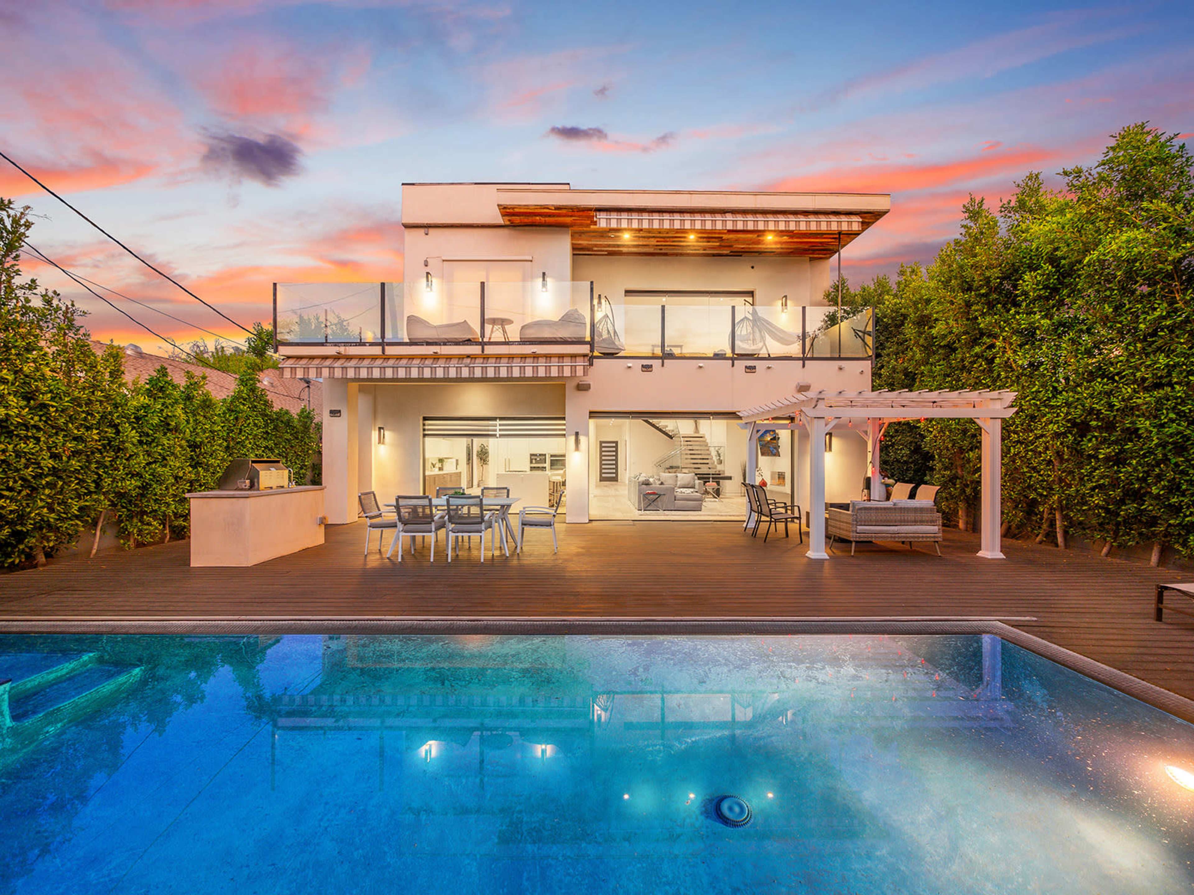 A modern two-story house with a swimming pool and deck overlooks a sunset sky filled with clouds.