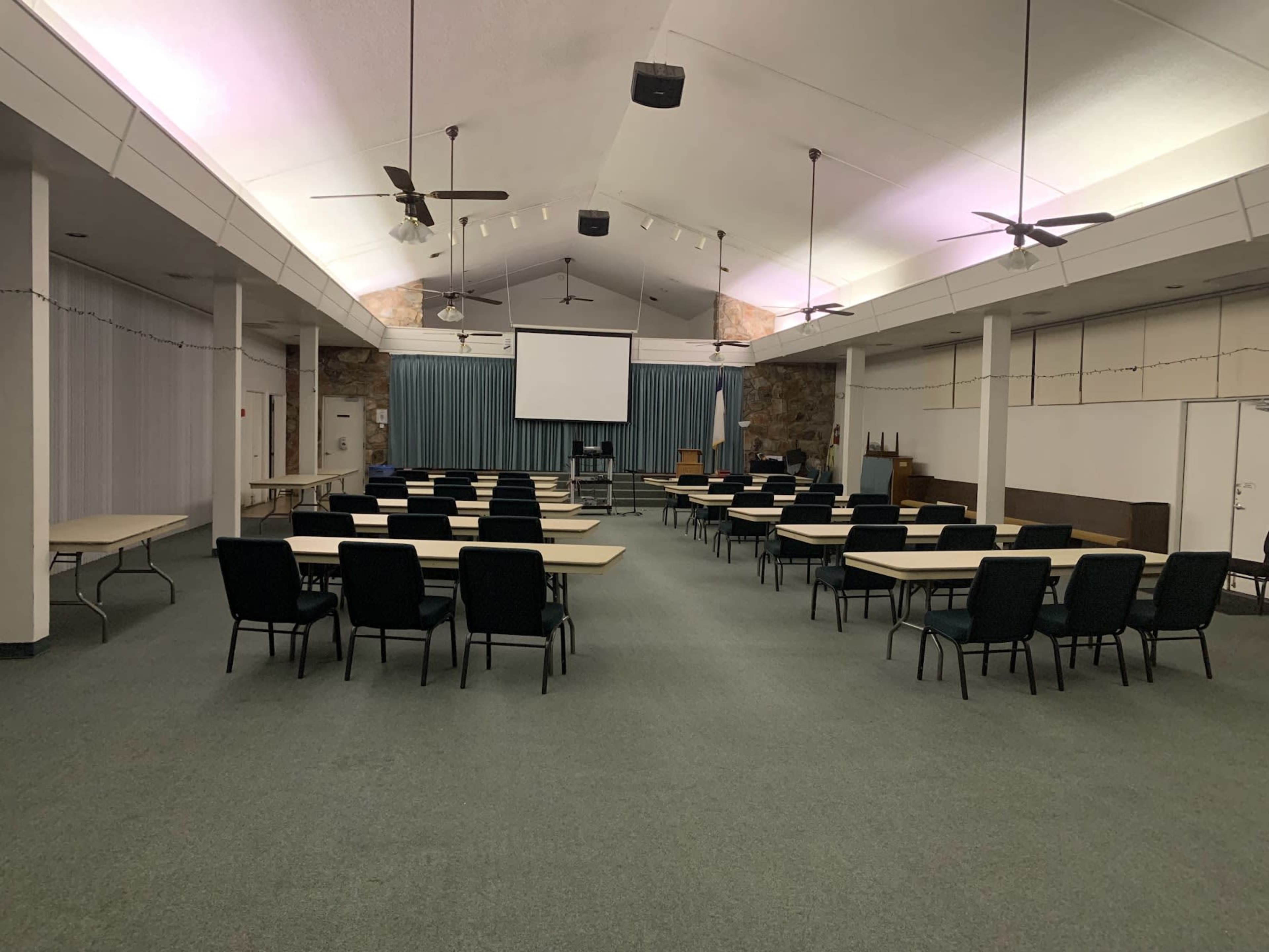 A multipurpose room set up with rows of tables and chairs facing a projector screen at the front.