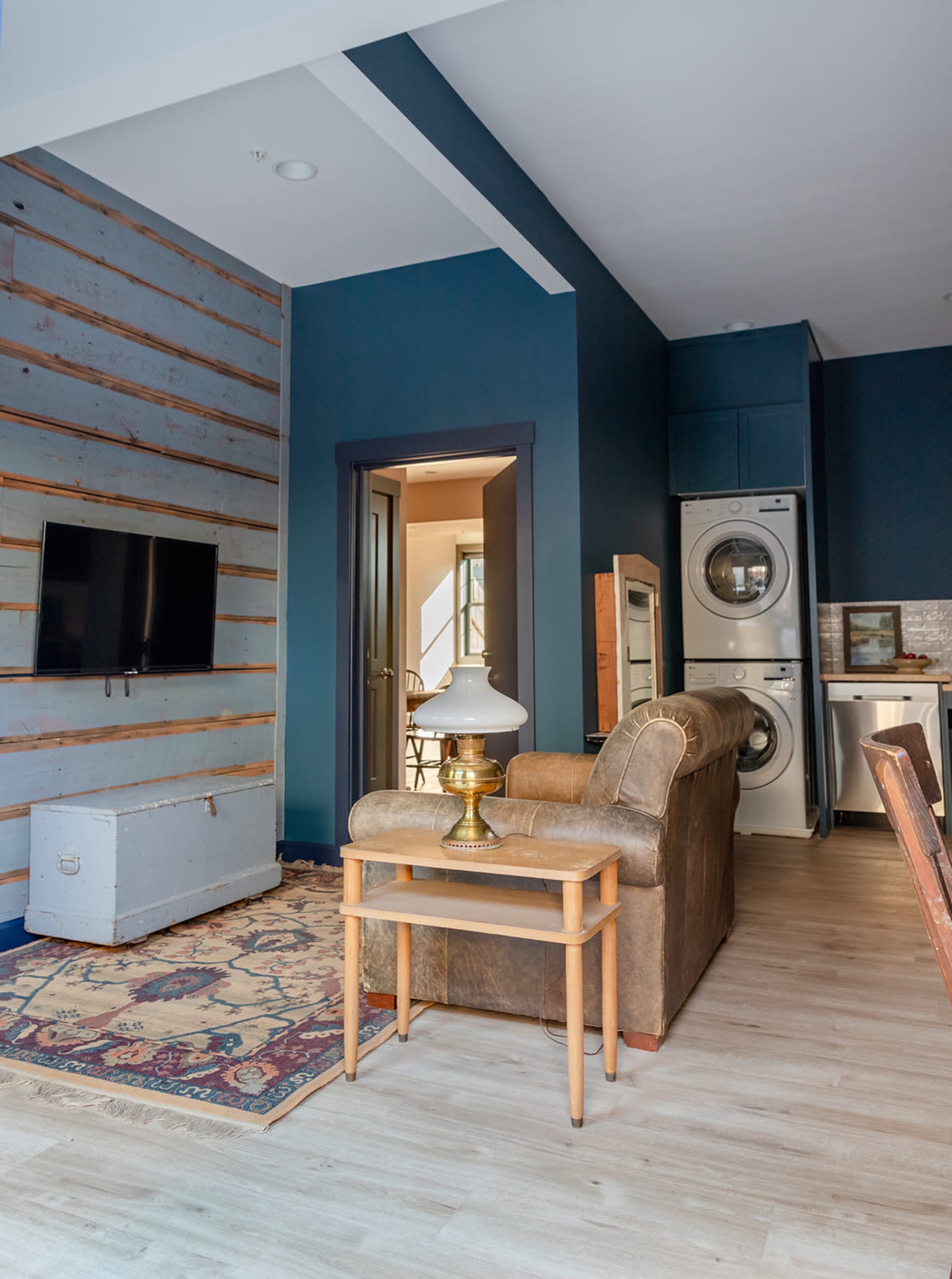 The image shows a modern living room with a brown sofa, a lamp on a wooden table, a television mounted on a textured wall, and laundry appliances visible in the background.
