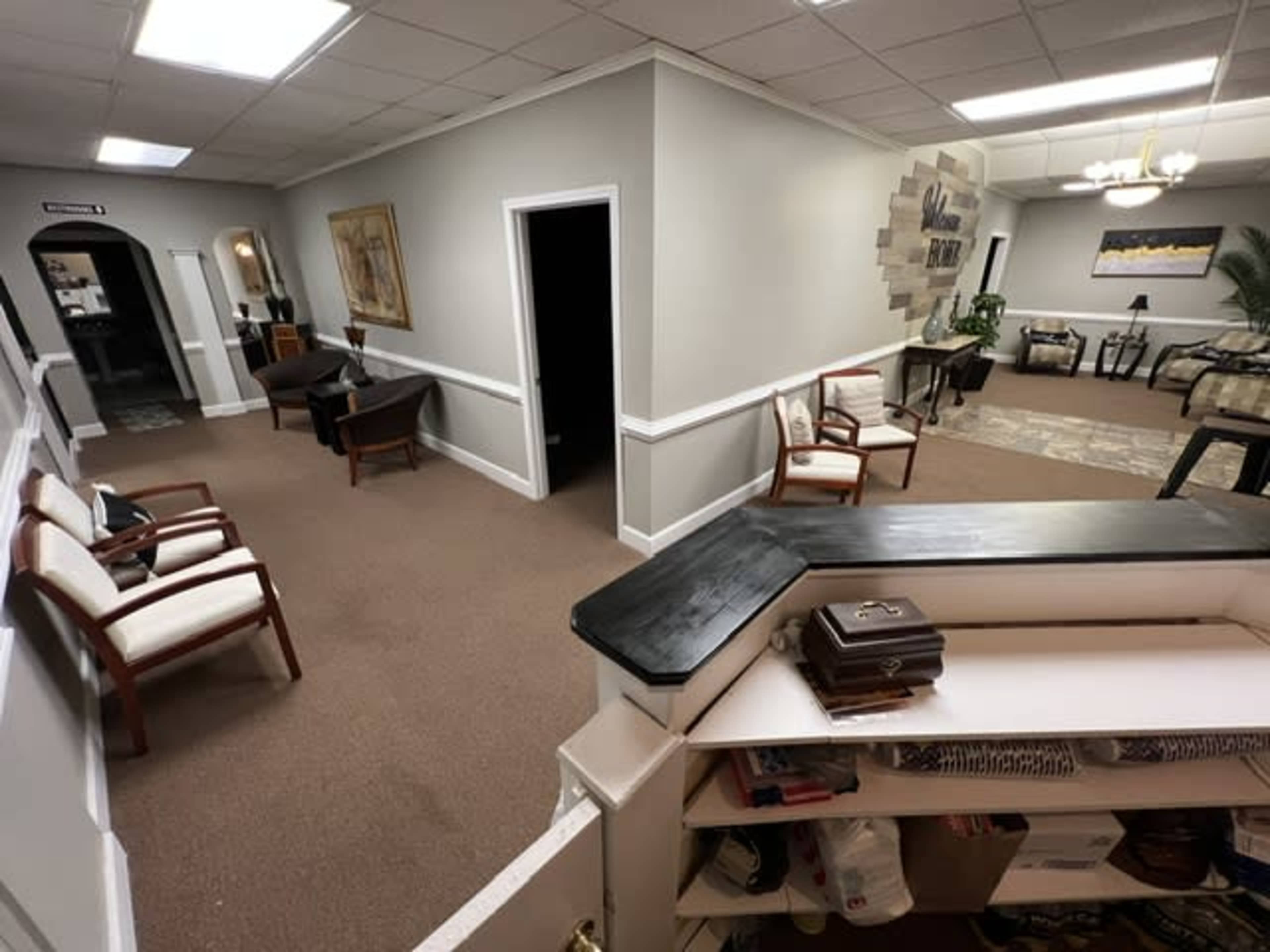 A corridor with chairs along the walls, leading to various rooms and a reception area with a desk.