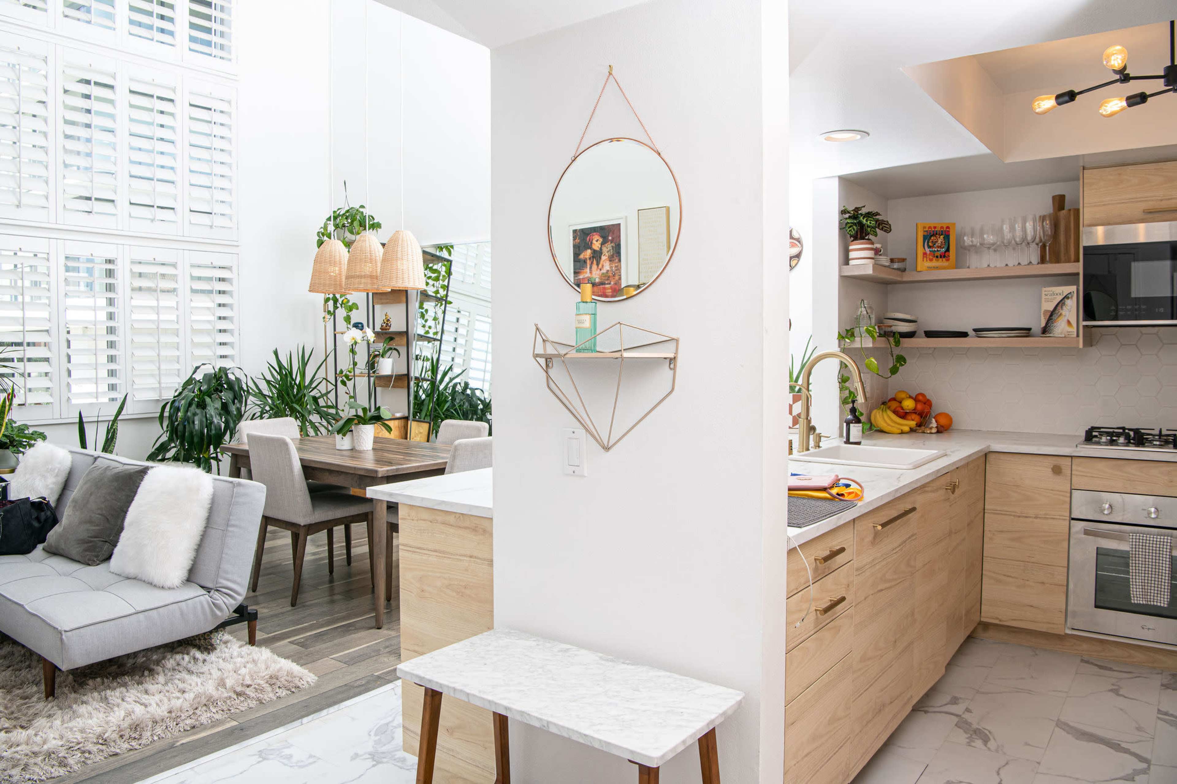 A modern kitchen and dining area with light wood cabinetry, a marble countertop, and large windows, featuring plants and a sitting area.