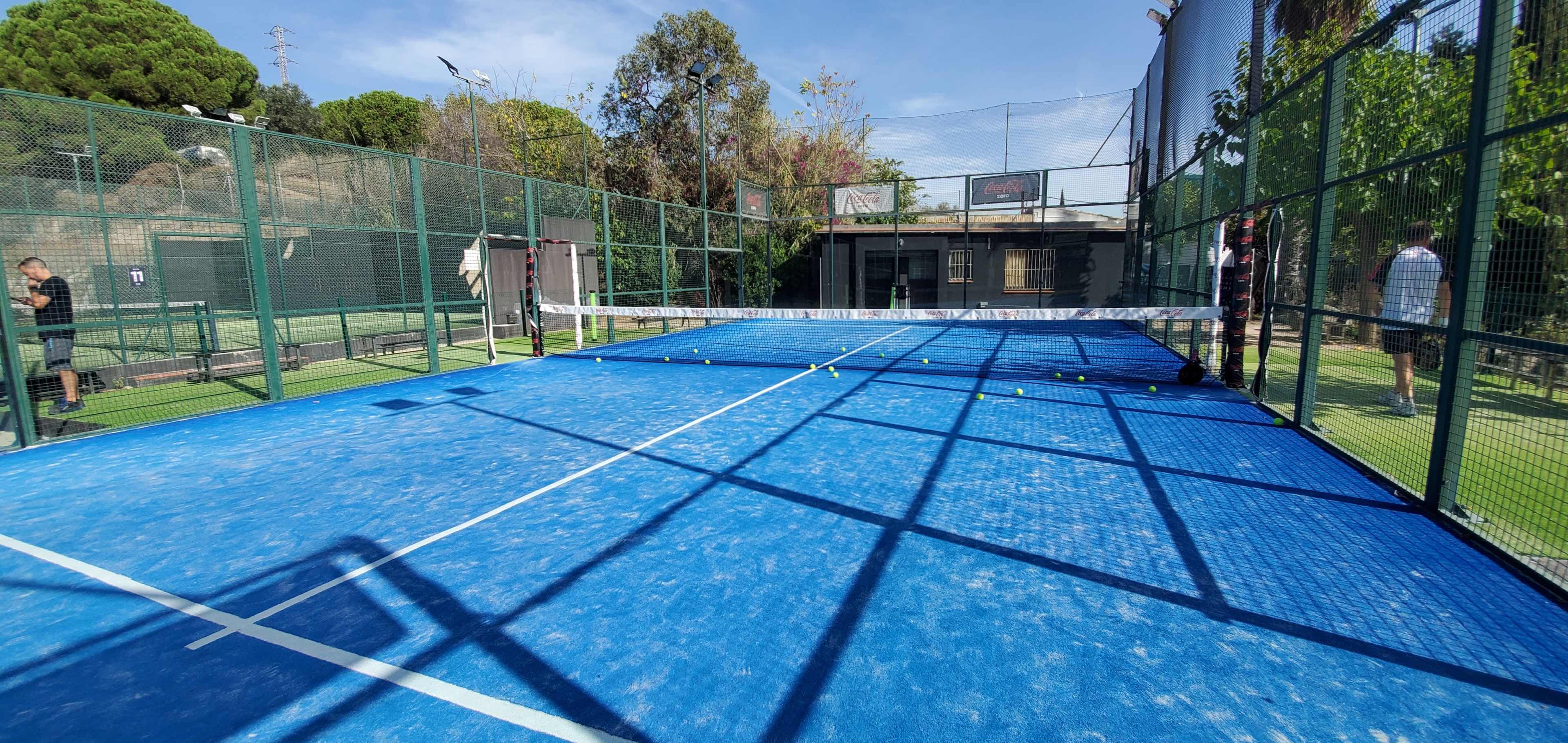 A blue paddle tennis court enclosed by fencing, with players on the sides and several balls scattered around the court.