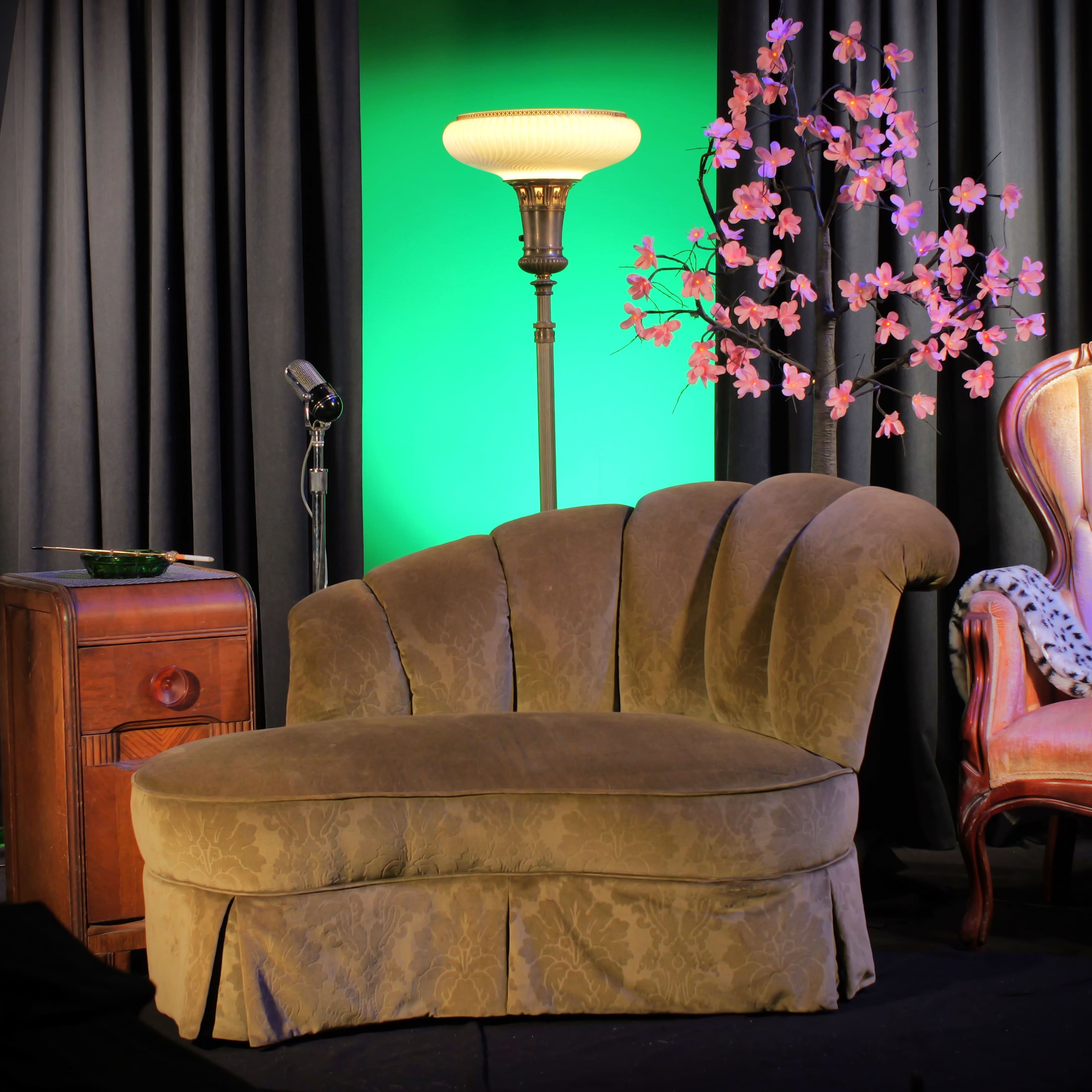 A vintage-style sofa with a curved backrest is positioned next to a standing lamp and a small wooden side table, set against a backdrop of dark curtains and pink-flowered decor.