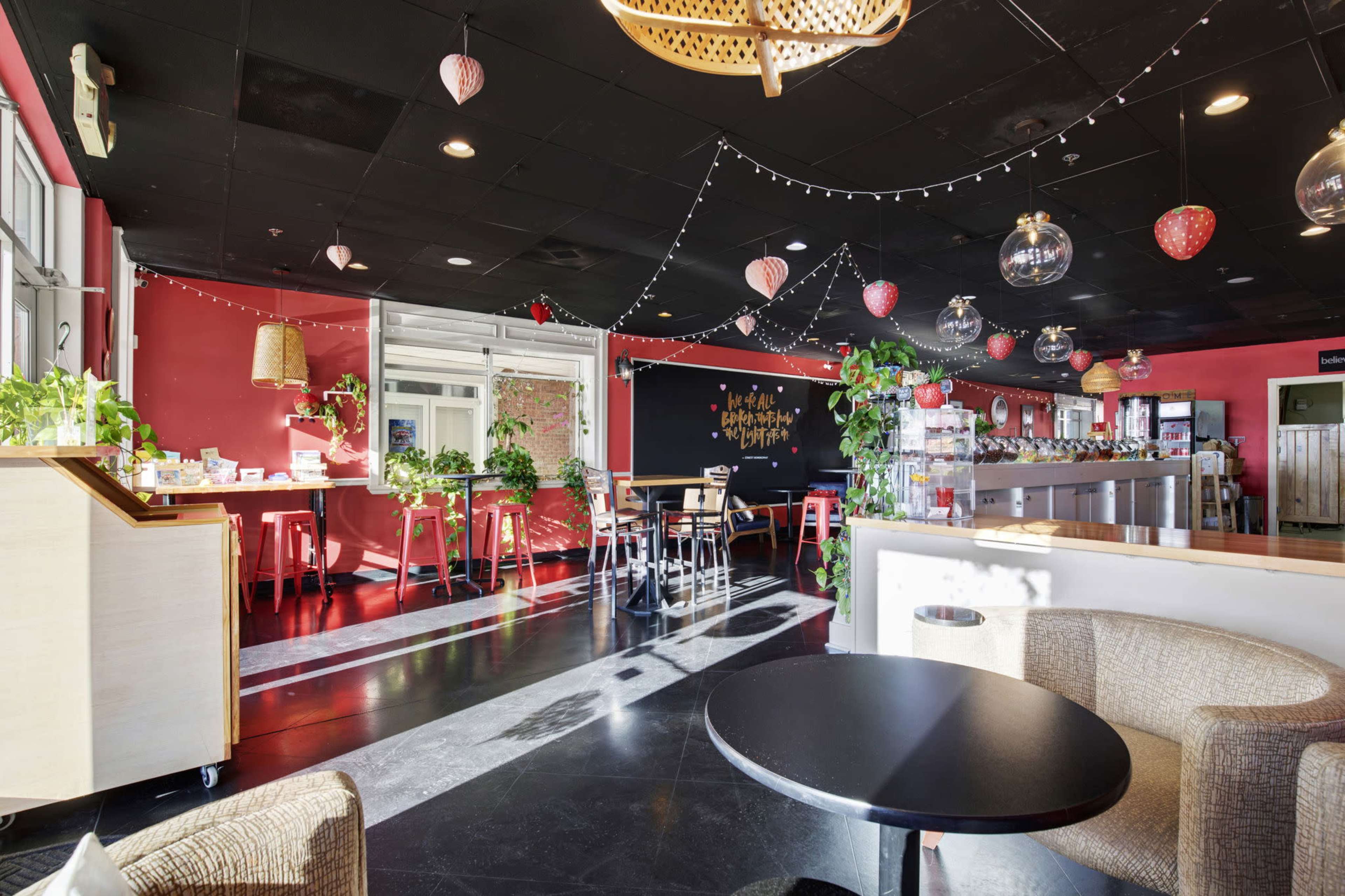 The image shows a brightly decorated café with red walls, hanging pendant lights, and plants scattered throughout the space, featuring a seating area with tables and chairs.