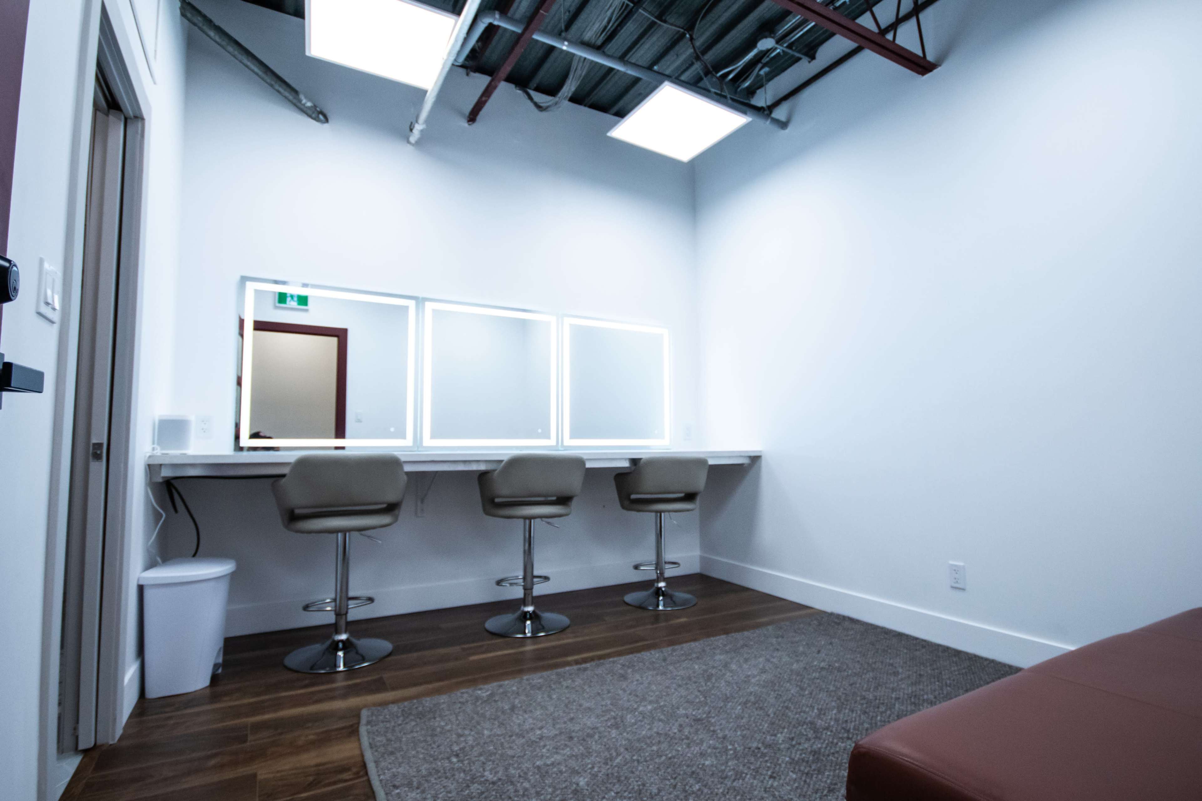 A minimalist makeup room featuring three bar stools at a long counter with three illuminated mirrors, a wastebasket, and a carpeted floor.