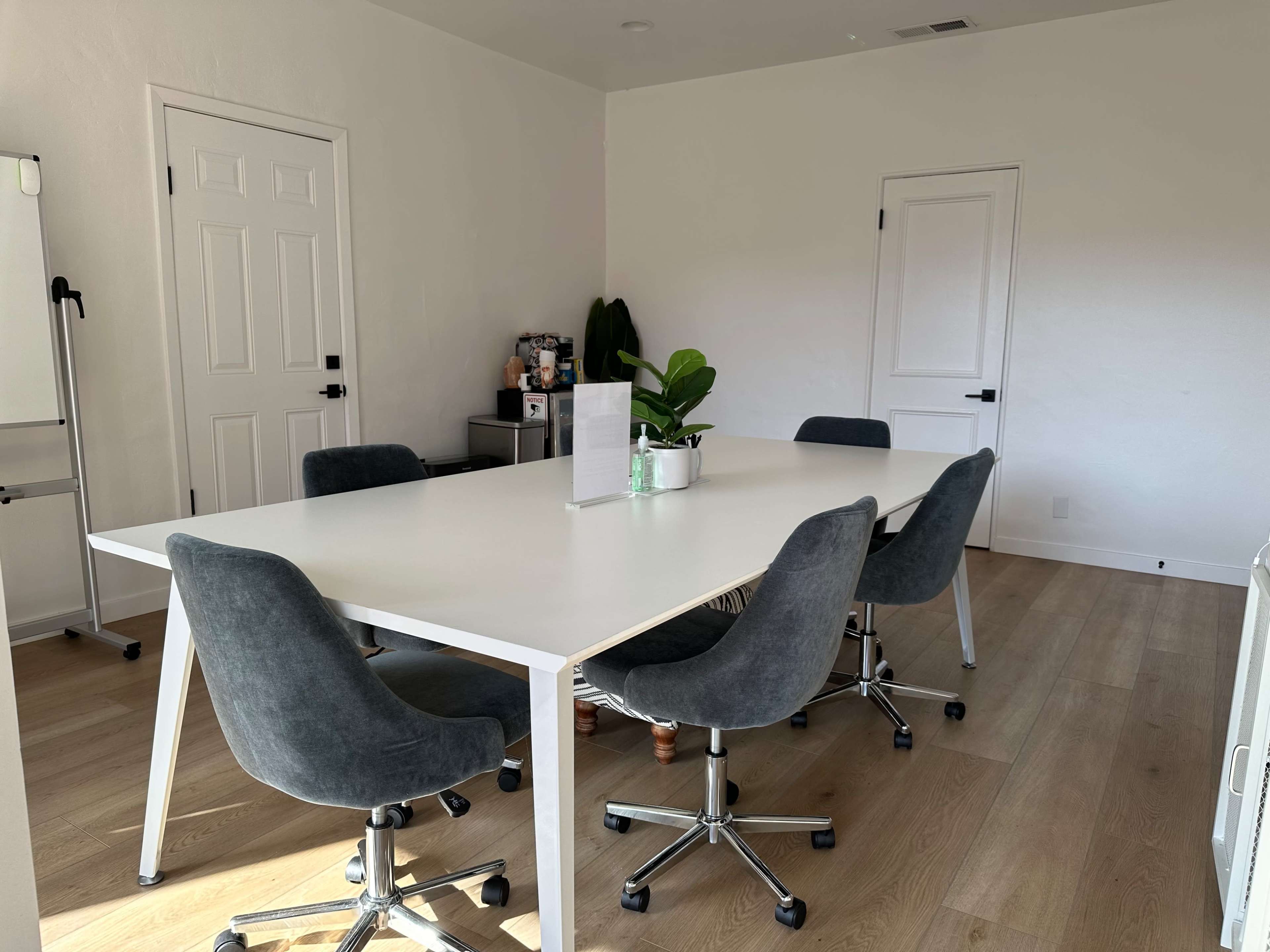 A modern conference room features a large white table with four gray chairs and a small plant on the table, set in a bright, minimalistic space.