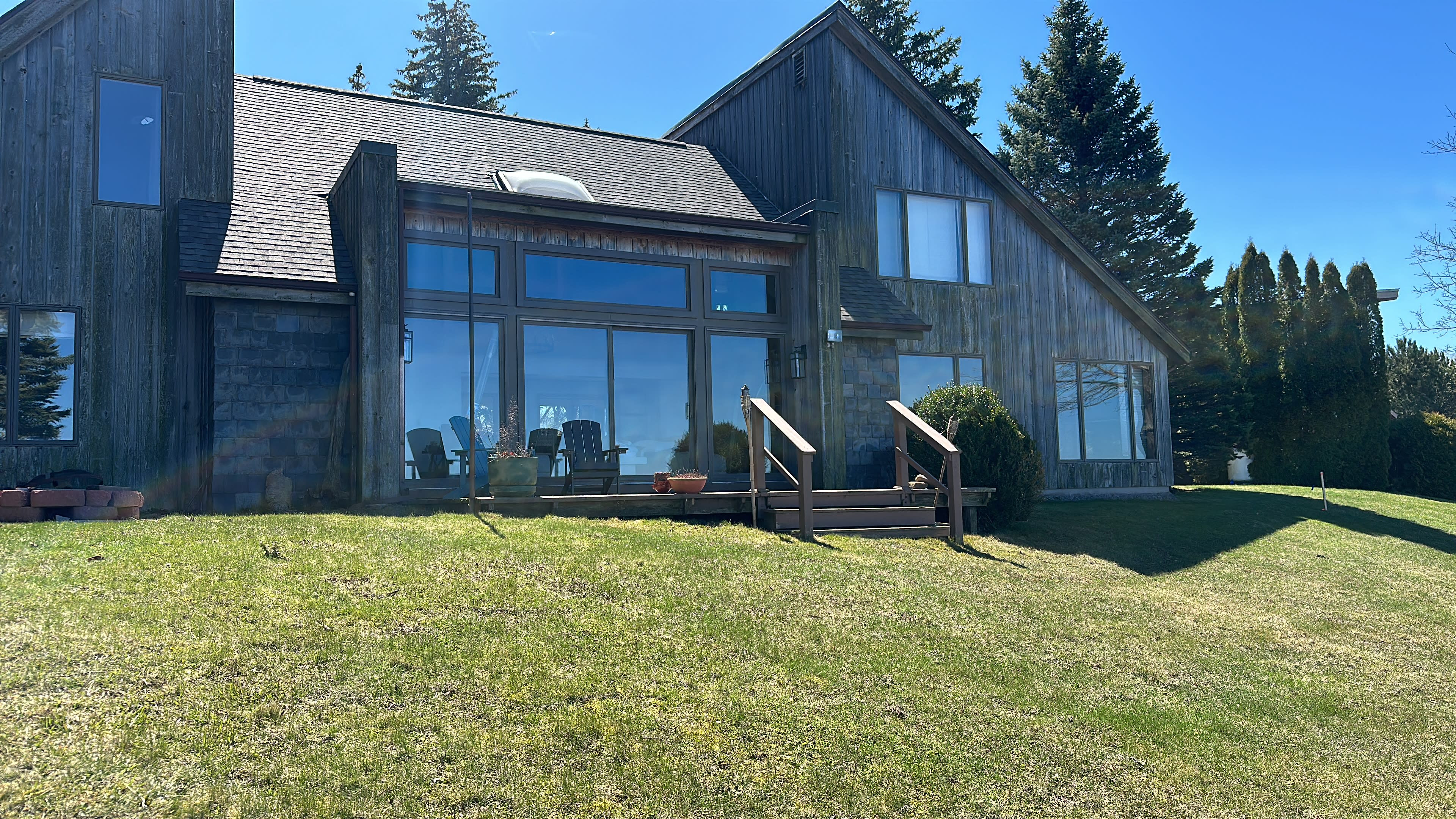 A large wooden house with large windows and a deck, set on a grassy lawn under a clear blue sky.