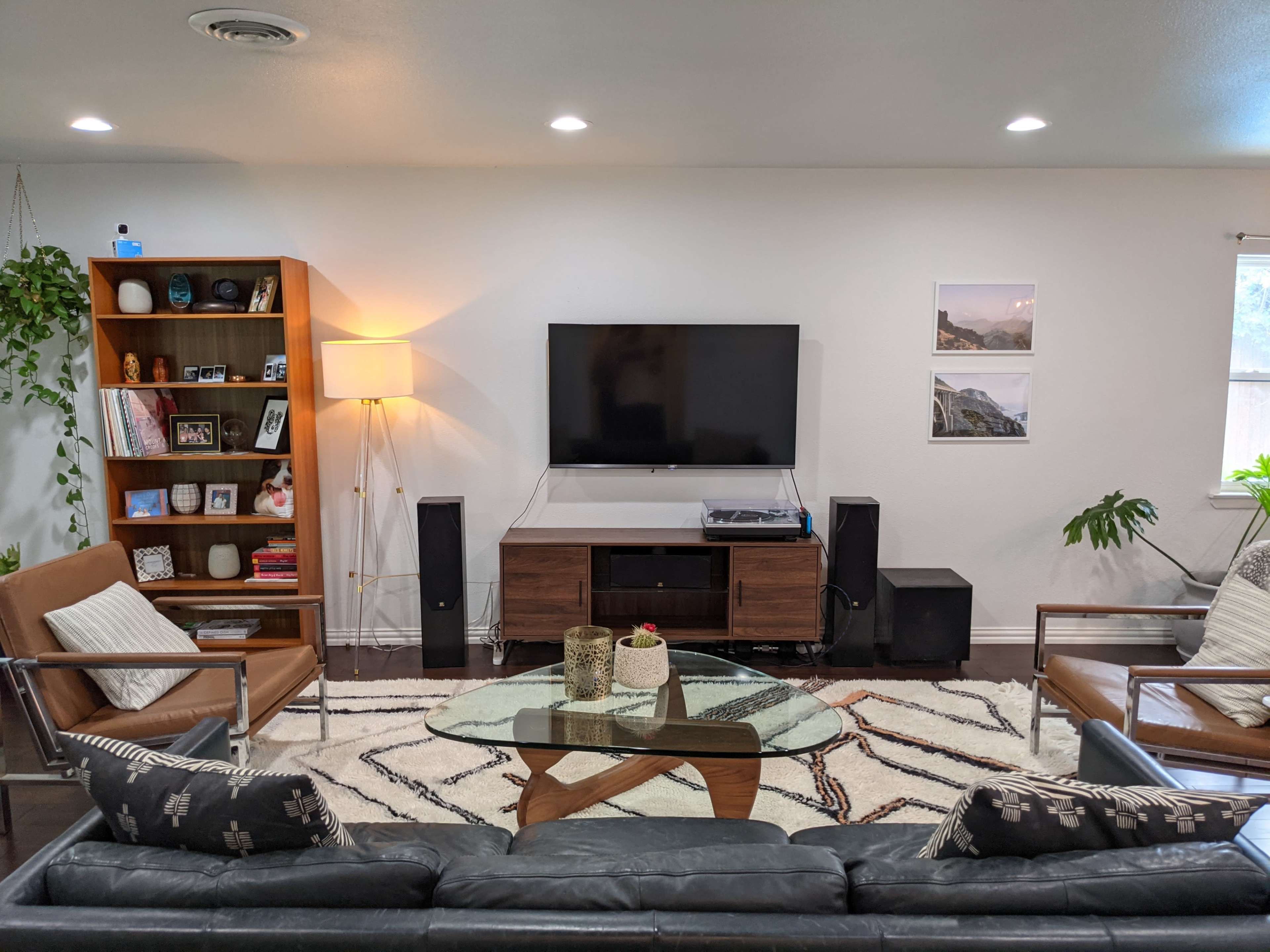 The living room features a dark leather sofa, wooden armchairs, a central coffee table, a television mounted on the wall, and various plants and decorative items.