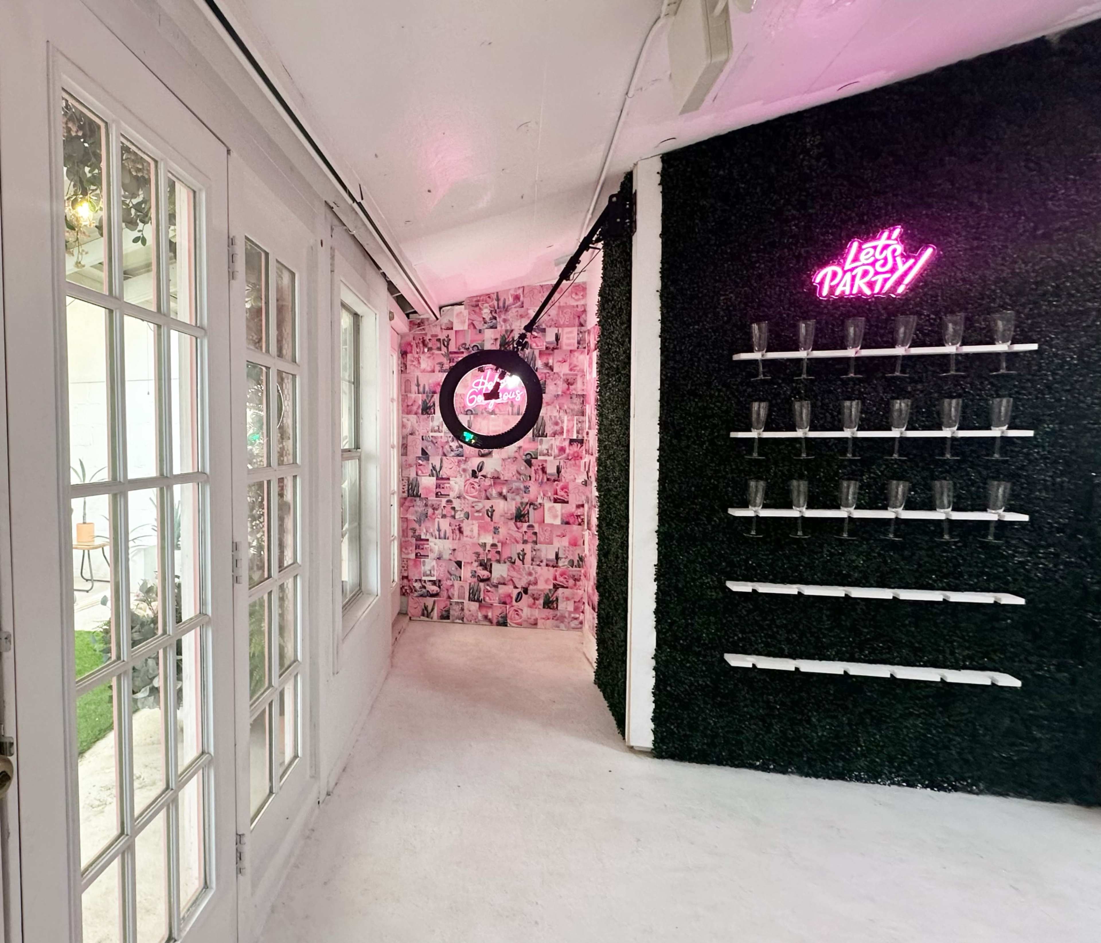 A brightly lit hallway with glass doors leading to a wall decorated with pink and black photo collages and a neon sign that reads "Let's PARTY!" next to shelves displaying clear glasses.