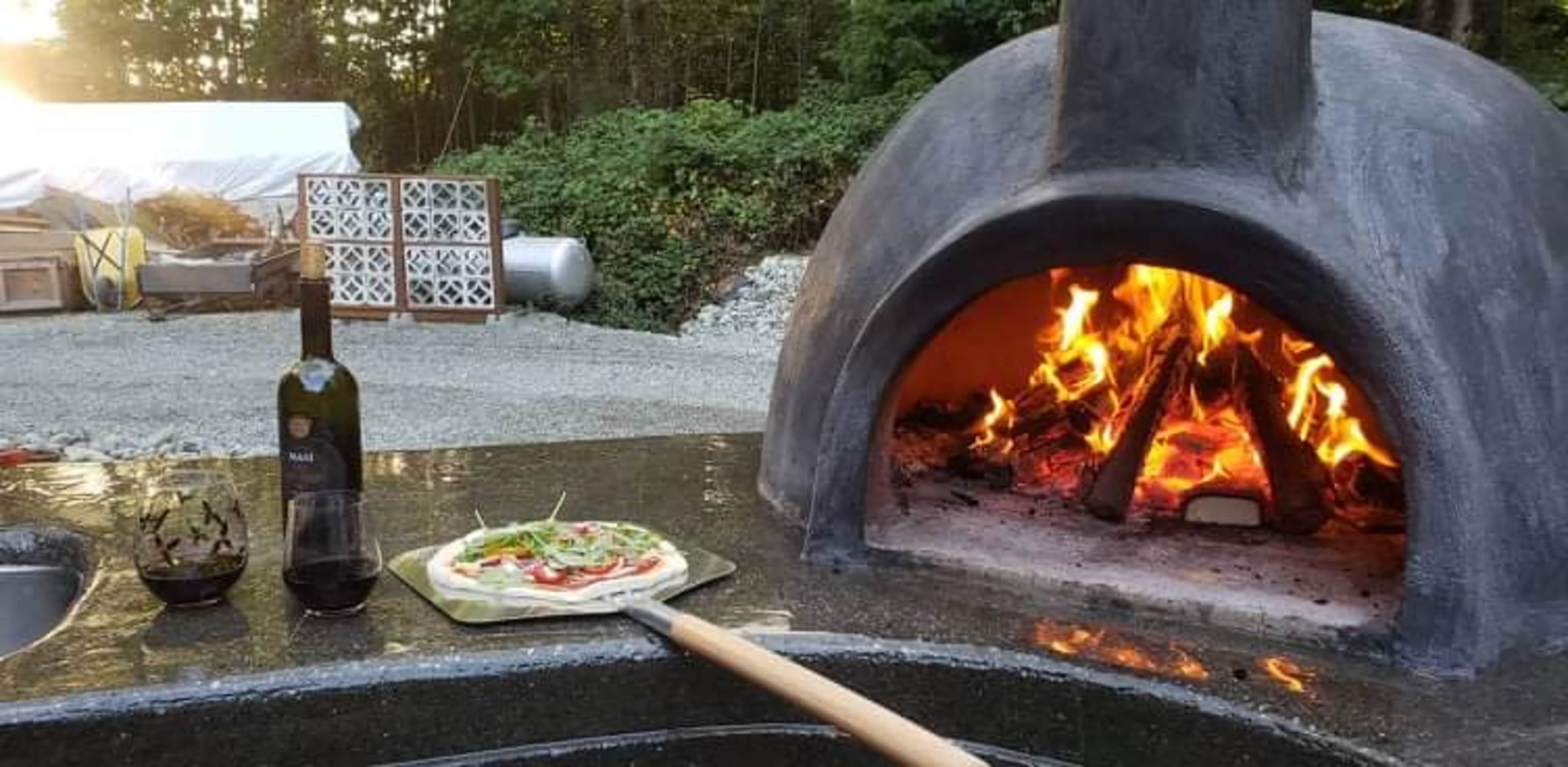 A pizza is being placed into a wood-fired oven with a bottle of olive oil and a glass of wine nearby.