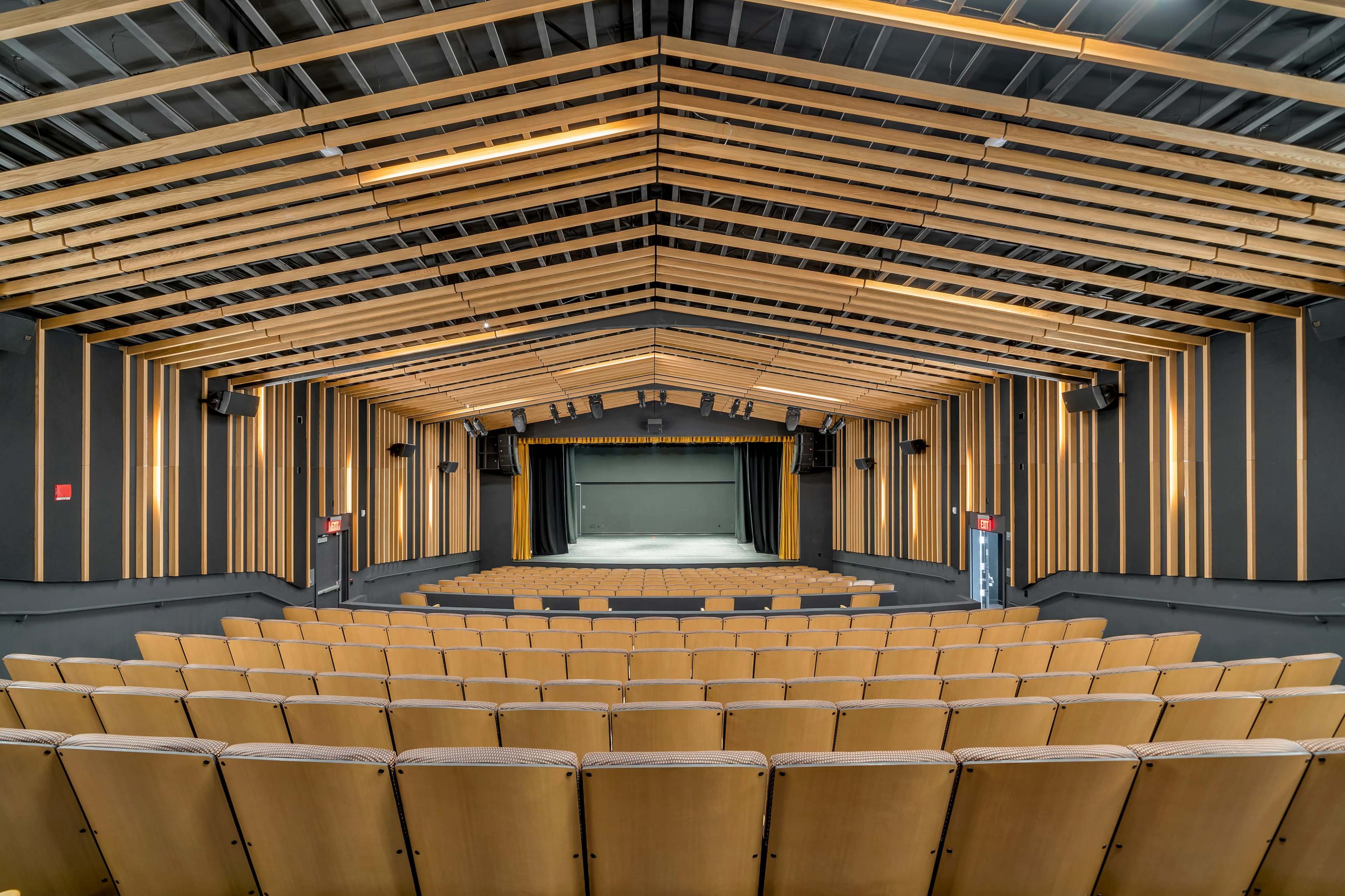 An empty auditorium with wooden seating arranged in a tiered formation facing a stage framed by a slanted ceiling and wooden beams.